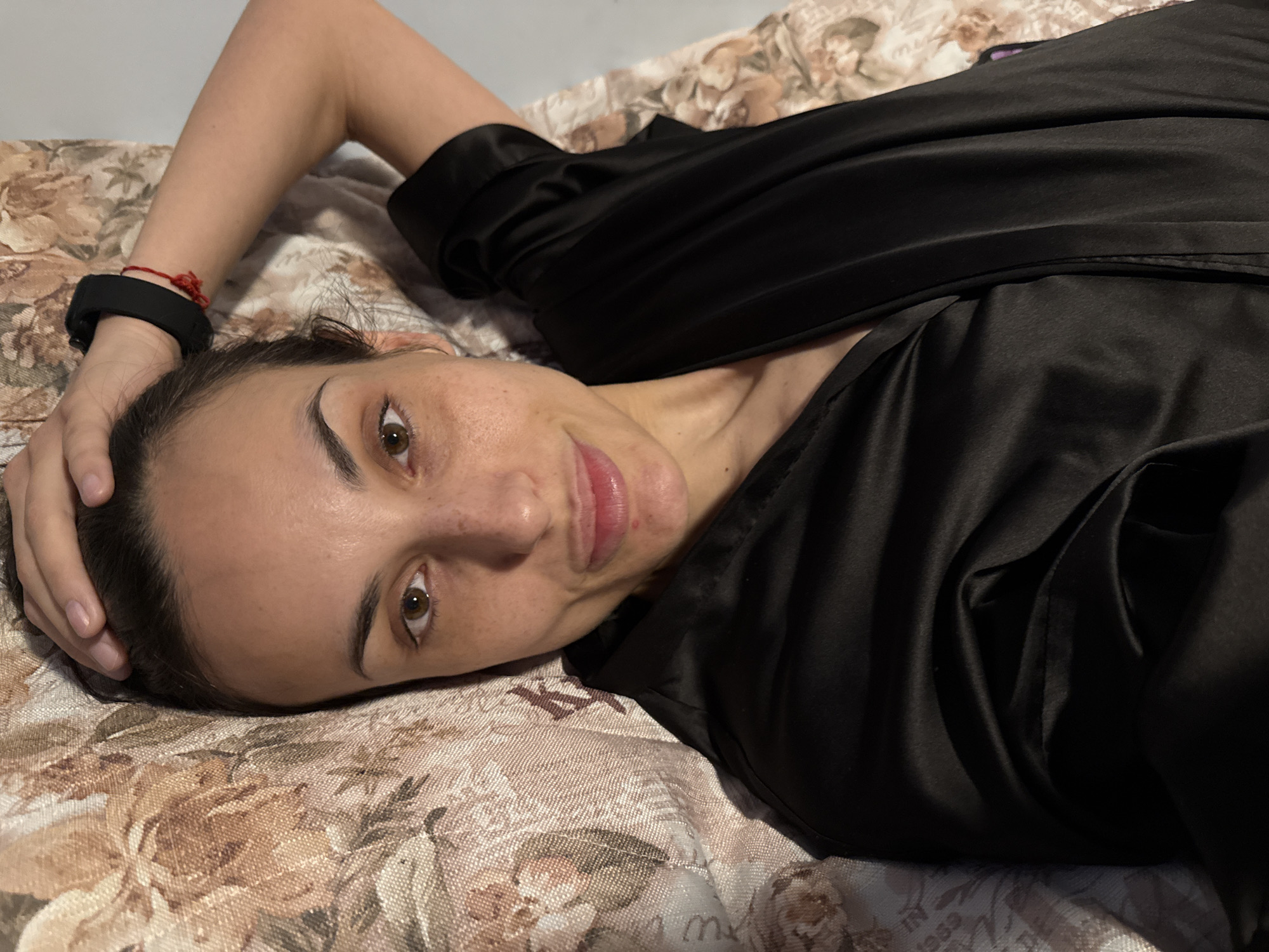 A person is lying on a bed with a floral patterned comforter. The person is facing the camera with their head tilted slightly to the left, and their left hand is positioned behind their head. They are wearing a black robe or garment. A black wristwatch and a red bracelet are visible on their left wrist. The person has brown eyes, dark hair, and freckles. The location in the city of Pleven, Bulgaria is not determined from the image.
