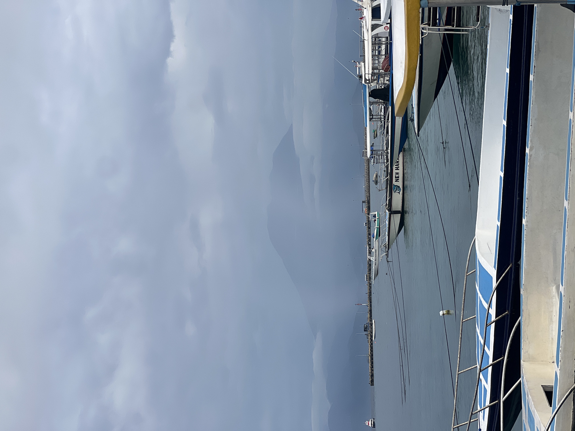 The provided image, rotated 90 degrees counter-clockwise, depicts a harbor scene in Nusa Penida, Indonesia, under a heavily overcast sky. When viewed correctly, the foreground and mid-ground show calm, dark water where several boats are docked.
On the immediate right side of the frame, a large vessel with a white hull featuring distinct blue and white striped patterns is prominently visible, equipped with a metal ladder. Further into the mid-ground, another boat displays the text "NEW HARBOUR" along its side, accompanied by a logo resembling a fish or dolphin. A long pier extends across the water in the distance, lined with smaller boats and sparse structures.
In the background, faint, layered silhouettes of large mountains or hills rise, blending into the expansive, uniform grey cloud cover that dominates the sky. The weather is heavily cloudy, indicating a dim daylight without direct sunlight. No specific human activity is discernible; the boats appear to be at rest in a quiet, working port environment.