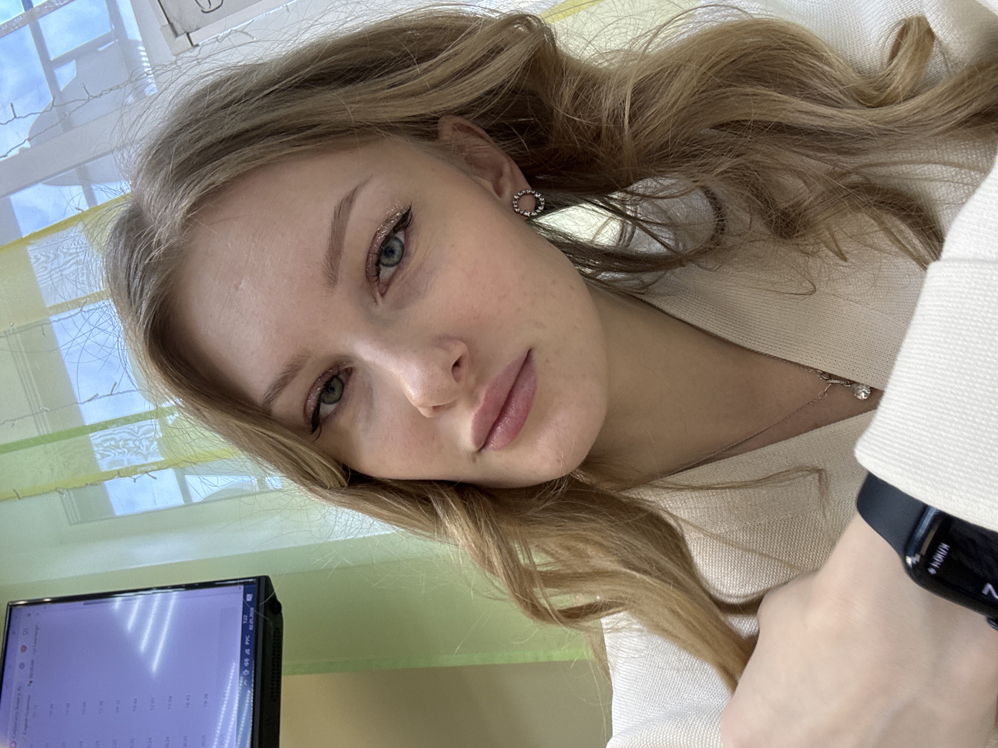 A fair-skinned young woman with light brown hair and green eyes is pictured in a selfie taken indoors, possibly at a workplace. She is wearing a light-colored blazer and a necklace with a small pendant. Her hair is styled in loose waves. She wears earrings and eye makeup. A smartwatch is visible on her wrist. A computer monitor is in the lower left corner of the image, and a window with blinds is in the background. Based on the location of the uploader, it is presumed that the image was taken in Perm, Russia.