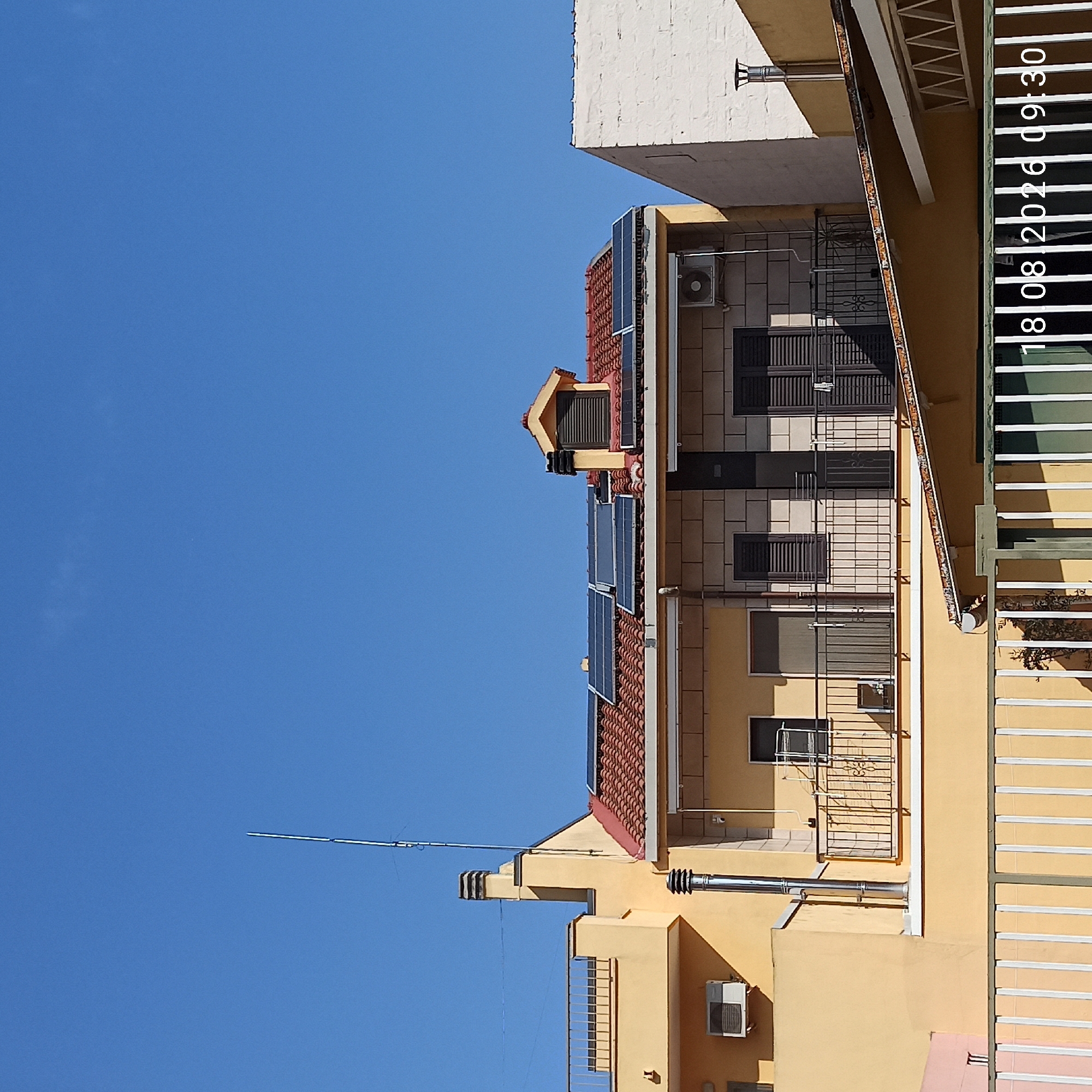 The image shows a bright, sunny day with a clear blue sky above a row of buildings. The foreground features a white metal fence. Behind the fence, a pale yellow building with a pink section on the far left is visible. This building has an air conditioning unit mounted on its exterior wall. Adjacent to it is another building with a red-tiled roof and several solar panels. This building also has a balcony with a black metal railing. A tall, slender antenna extends vertically from the top of the yellow building. Further to the right, a white building with a red-tiled roof and a chimney is present. On the right side of the image, a large section of a white building is visible. A timestamp at the bottom right corner of the image reads "18 08 2026 09:30".