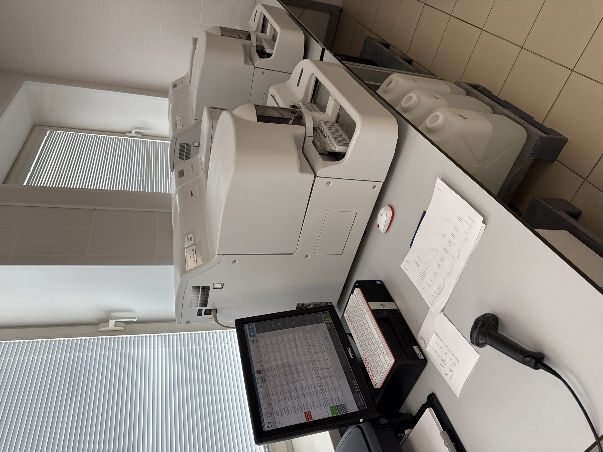 The image shows a laboratory setting, likely in Moscow, Russia, based on the Cyrillic script on the screen and papers. Three automated laboratory analyzers are arranged along a countertop, with a computer workstation in the foreground. The workstation includes a monitor displaying a table of data, a compact keyboard, a tower computer, and a barcode scanner. Several sheets of paper with graphs and text are scattered on the counter. A window with closed blinds is visible to the left, providing natural light. Containers of reagent or waste solutions are positioned behind the analyzers. The environment appears clean and well-lit, suggesting a modern laboratory facility. No people are visible in the frame.