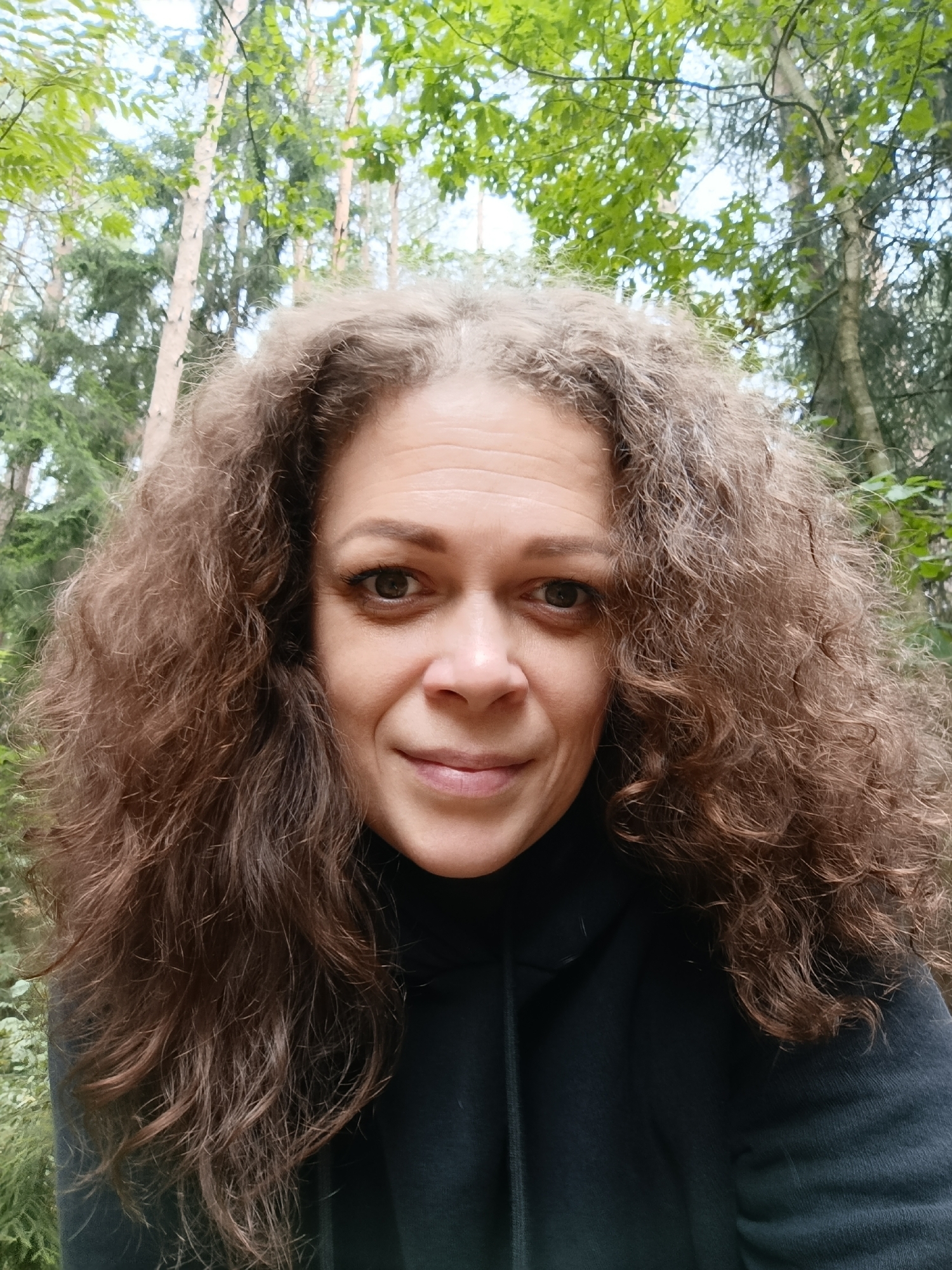 A medium shot, selfie-style, captures a woman with curly brown hair from the chest up, outdoors. She is wearing a black hooded sweatshirt. Her face is angled slightly to the left, and she is looking directly at the camera with a faint smile. Her hair is voluminous and frames her face. The background consists of trees with green foliage and visible branches, suggesting a forest or wooded area. The lighting appears natural, indicating it is daytime. The woman's visible skin tone is light.