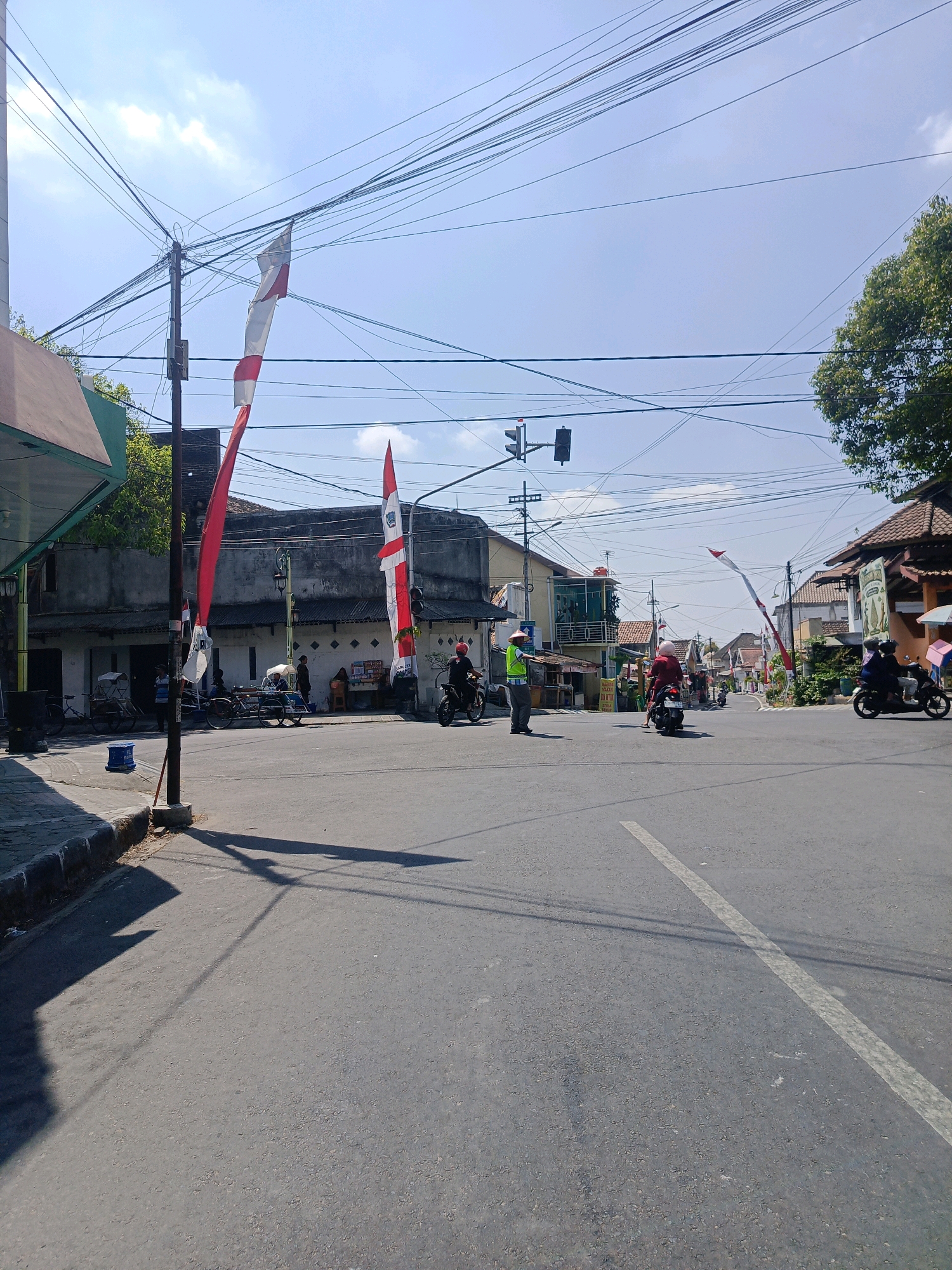 The image captures a street intersection in Kebonagung, Indonesia, on a clear day. The foreground shows a paved road with a white lane marker. On the left side of the road, a sidewalk with a curb is visible, leading to a building with a green awning and a concrete wall. A utility pole with a dense network of wires stands on the sidewalk, adorned with two Indonesian flags, one partially obscured.
The intersection is active with several people and vehicles. Two men in reflective vests are directing traffic, one standing near the utility pole and another further down the road. Several motorcycles are present, some carrying passengers. A few people are seated on bicycles and what appear to be pedicabs. Buildings line both sides of the street, displaying a mix of architectural styles, including traditional tiled roofs. The general ambiance is that of a typical urban street scene in Indonesia, with ample overhead electrical wiring and flags suggesting a local celebration or national holiday. The lighting indicates it is daytime, with shadows cast by the buildings and utility pole.