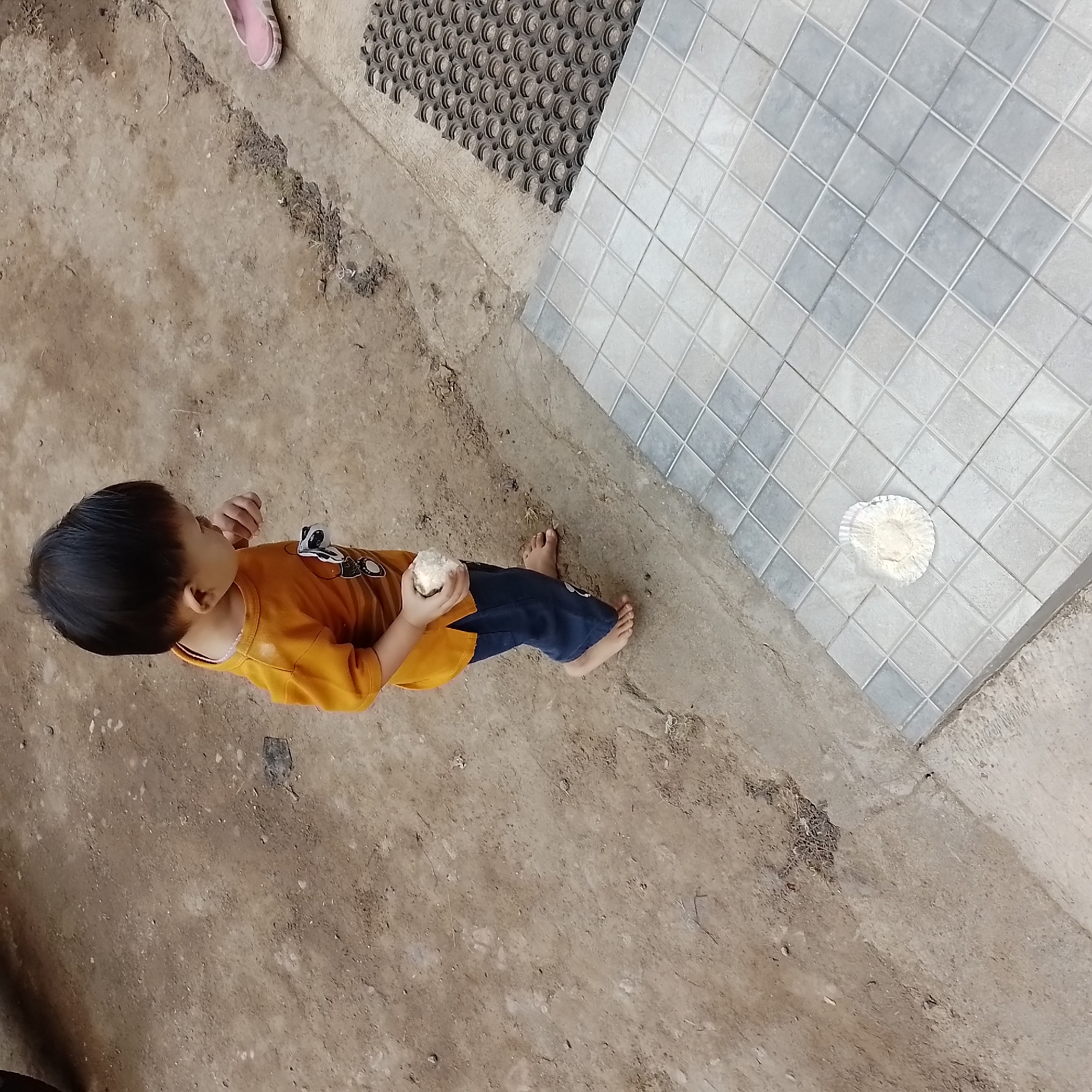 A young child, with dark hair and wearing an orange t-shirt and blue pants, stands barefoot on a dirt surface. The child is holding a light-colored object in their right hand and appears to be looking down at it. In the foreground, a tiled patio is visible, with a small pile of light-colored powder on one of the tiles. A black rubber mat is also present on the patio, and a pink flip-flop is on the dirt to the right. The background consists of a rough, unpaved area with some scattered debris. The overall impression is an outdoor scene, likely a residential yard or pathway.