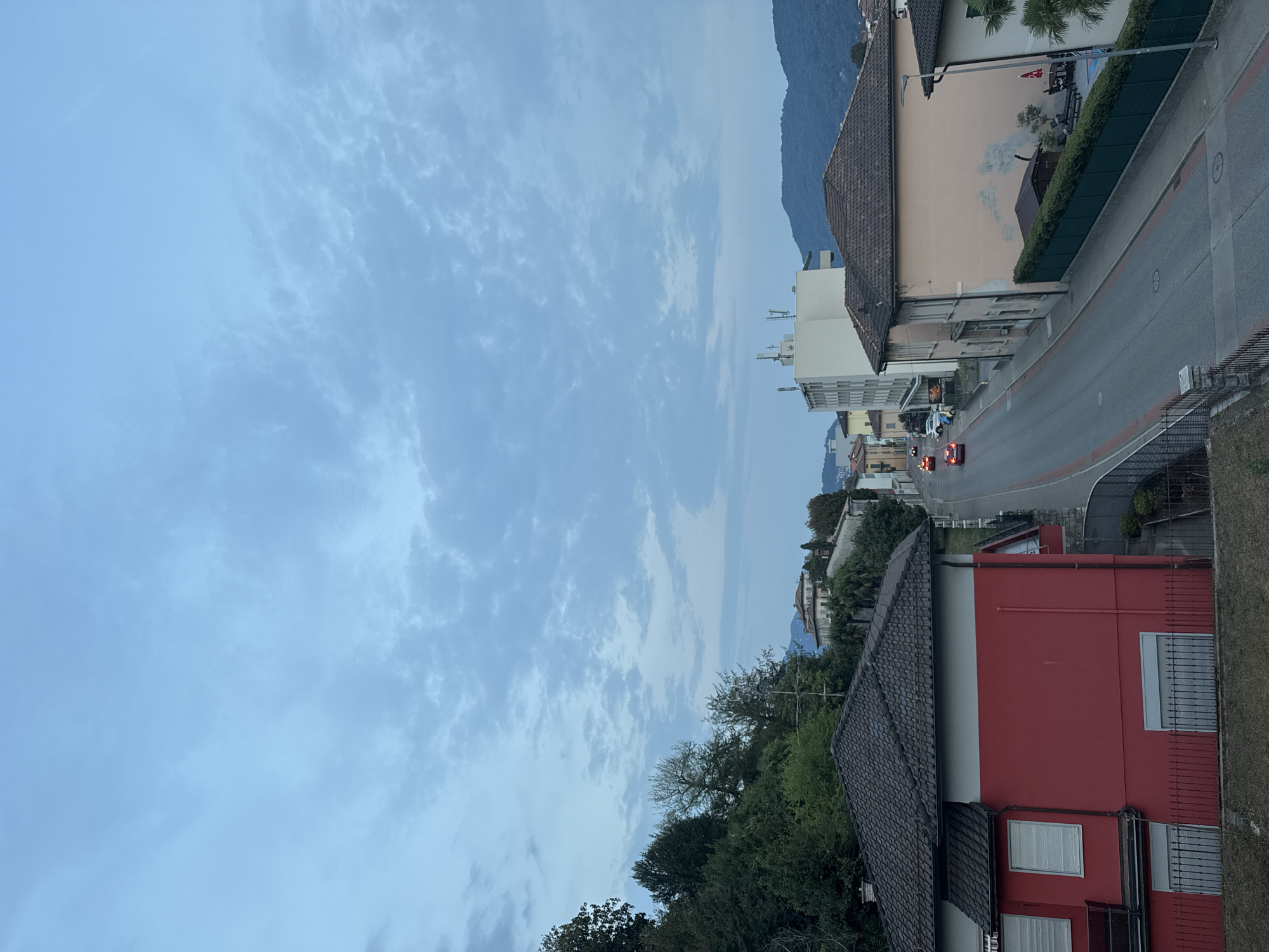 An outdoor, high-angle, eye-level, wide shot captures a suburban street in Balerna, Switzerland, under a cloudy sky. The road, bordered by red lines, curves through the scene, with a few cars driving on it. On the left, a red house with white window shutters and a brown tiled roof is prominent. Across the street, a light-colored building with a white facade and multiple windows stands tall. A light pink building with a brown tiled roof is also visible. Lush green trees and vegetation cover a hillside on the left, while mountains form a backdrop on the right. The time appears to be dusk or early morning due to the soft lighting and muted colors.