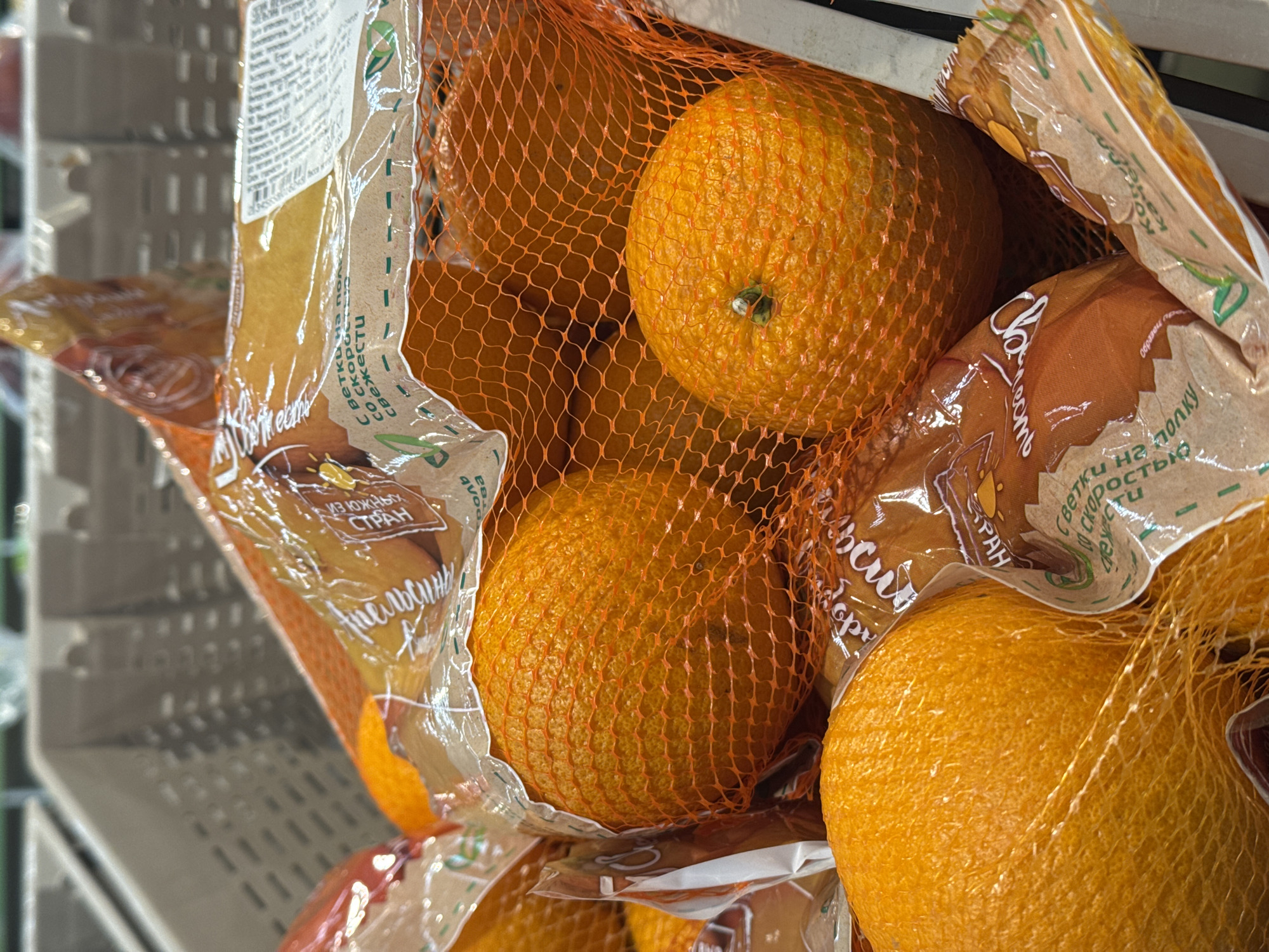 This is a close-up shot of a bag of oranges. The oranges are in an orange mesh bag, and the bag appears to be made of plastic. Some labeling on the bag is in Russian. The oranges are stacked in what appears to be a plastic crate. Based on the labeling, the oranges are likely sourced from southern countries and available on shelves quickly. No specific information ties this image to Ново-Никольское, Russia, making it an assumption.