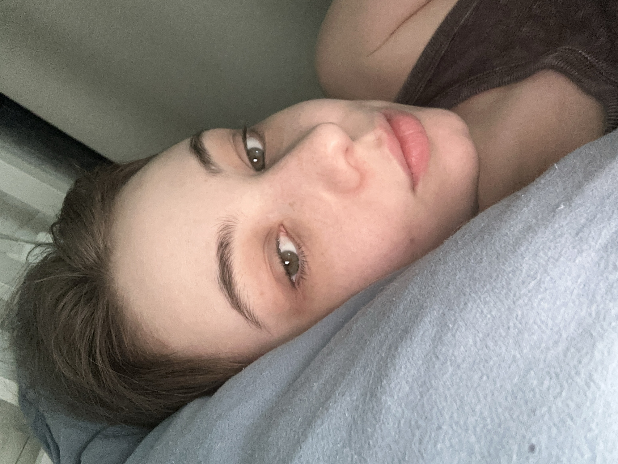 A close-up, angled shot depicts a person lying down on a light blue pillow, possibly in a domestic setting. The person's face is the main focus. They have fair skin, with visible freckles, and light brown hair. Their eyes are open and appear to be hazel or light brown. The person is wearing a brown tank top. A black, horizontal object is visible in the background near the top left of the frame. The exact location within Moscow, Russia cannot be determined from the visual information provided.