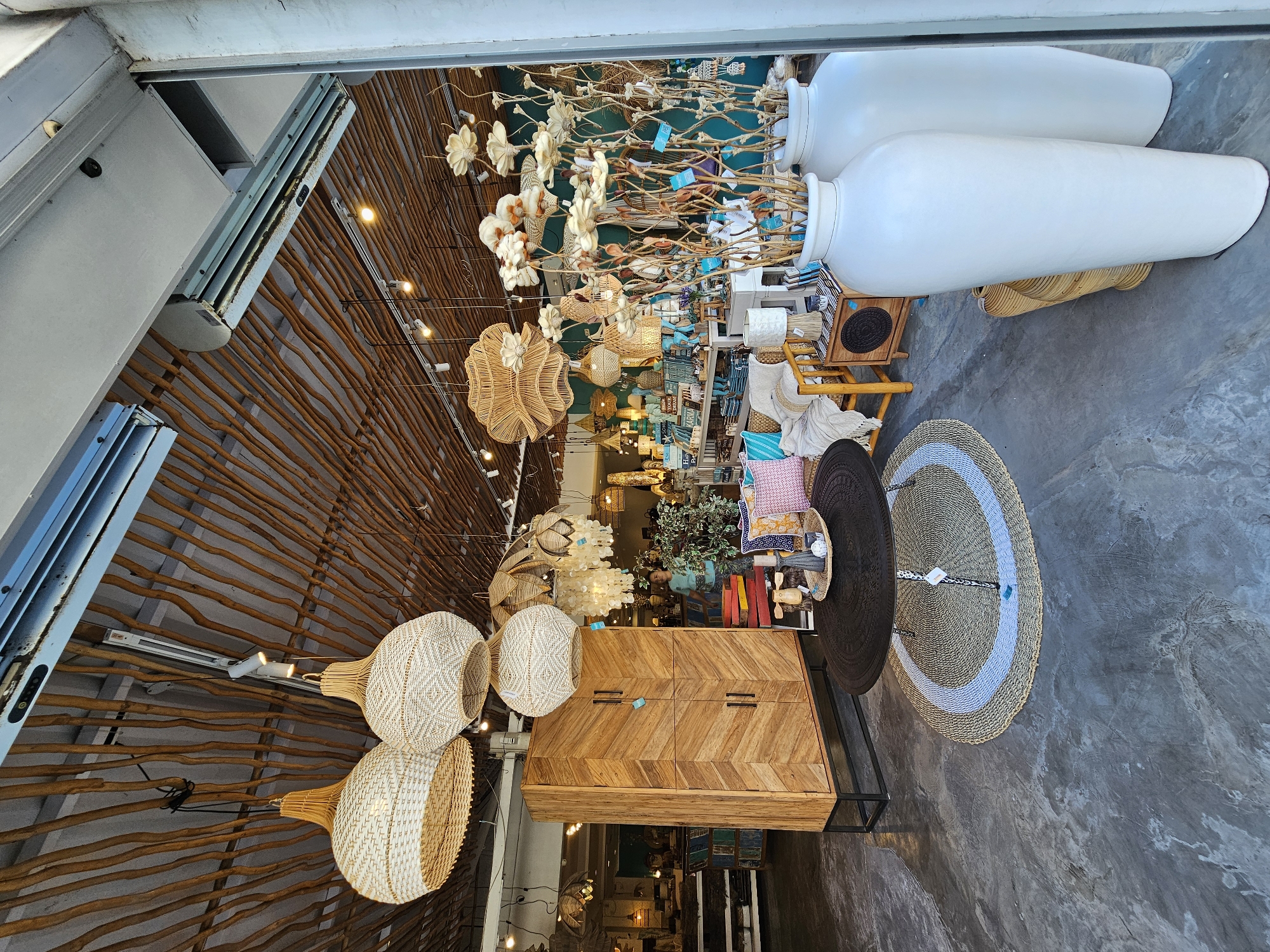The image depicts the interior of a retail store, possibly a home decor or gift shop, located in Denpasar, Indonesia. The ceiling is constructed with natural wooden branches, and several woven pendant lights hang from it. In the foreground, a dark, intricately carved round coffee table rests on a circular woven rug with a light blue stripe. Behind the table, a tall, wooden cabinet with a chevron pattern and black handles stands on a black metal base.
To the right of the cabinet, there is a wooden chair with a woven seat and back, adorned with throw pillows. A side table next to the chair holds a lamp and decorative items. Further to the right, large white vases are displayed, one of which contains dried floral arrangements with light-colored, papery flowers and natural branches.
The store's interior features teal-colored walls in the background, where more woven light fixtures, shelves with merchandise, and other decorative items are visible. A person, appearing to be a woman wearing a light blue shirt, is partially visible in the background, browsing the merchandise. The floor is a polished concrete surface with visible variations in tone and texture. The overall ambiance suggests a bohemian or naturalistic aesthetic.
