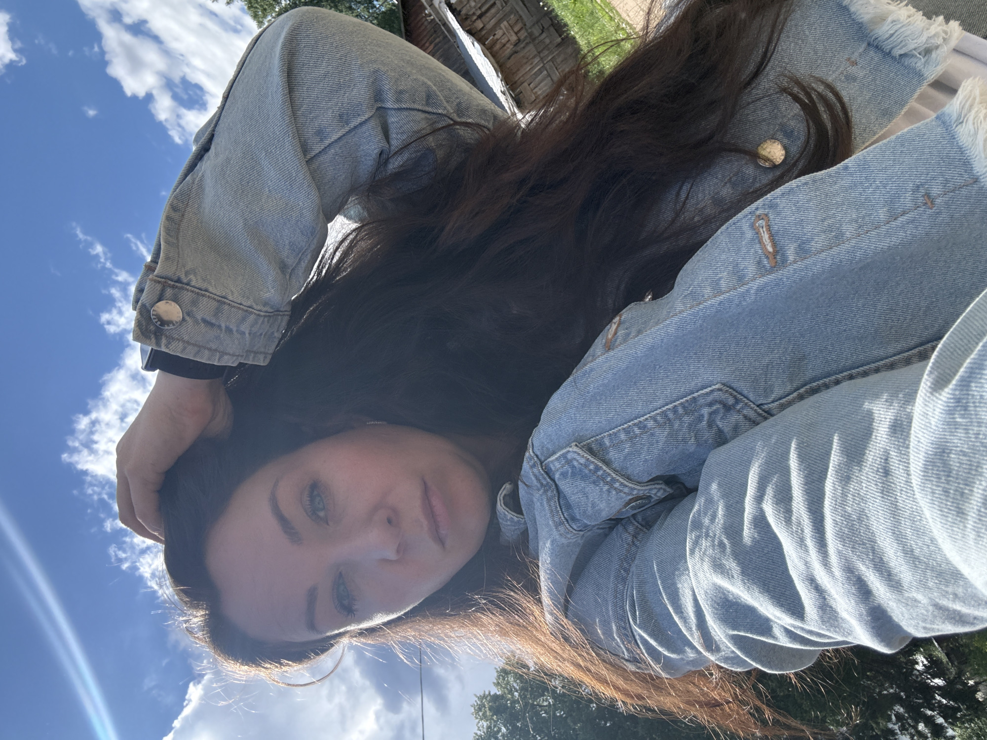 The image shows a woman from a low-angle perspective. She is wearing a light blue denim jacket. Her dark brown hair is partially covering her face. She is holding her hair with her hand. The sky is visible in the background with scattered clouds. The location is possibly a park in Chaikovsky, Russia, as indicated by the presence of trees and greenery in the background. The angle suggests the photo was taken by the subject herself.
