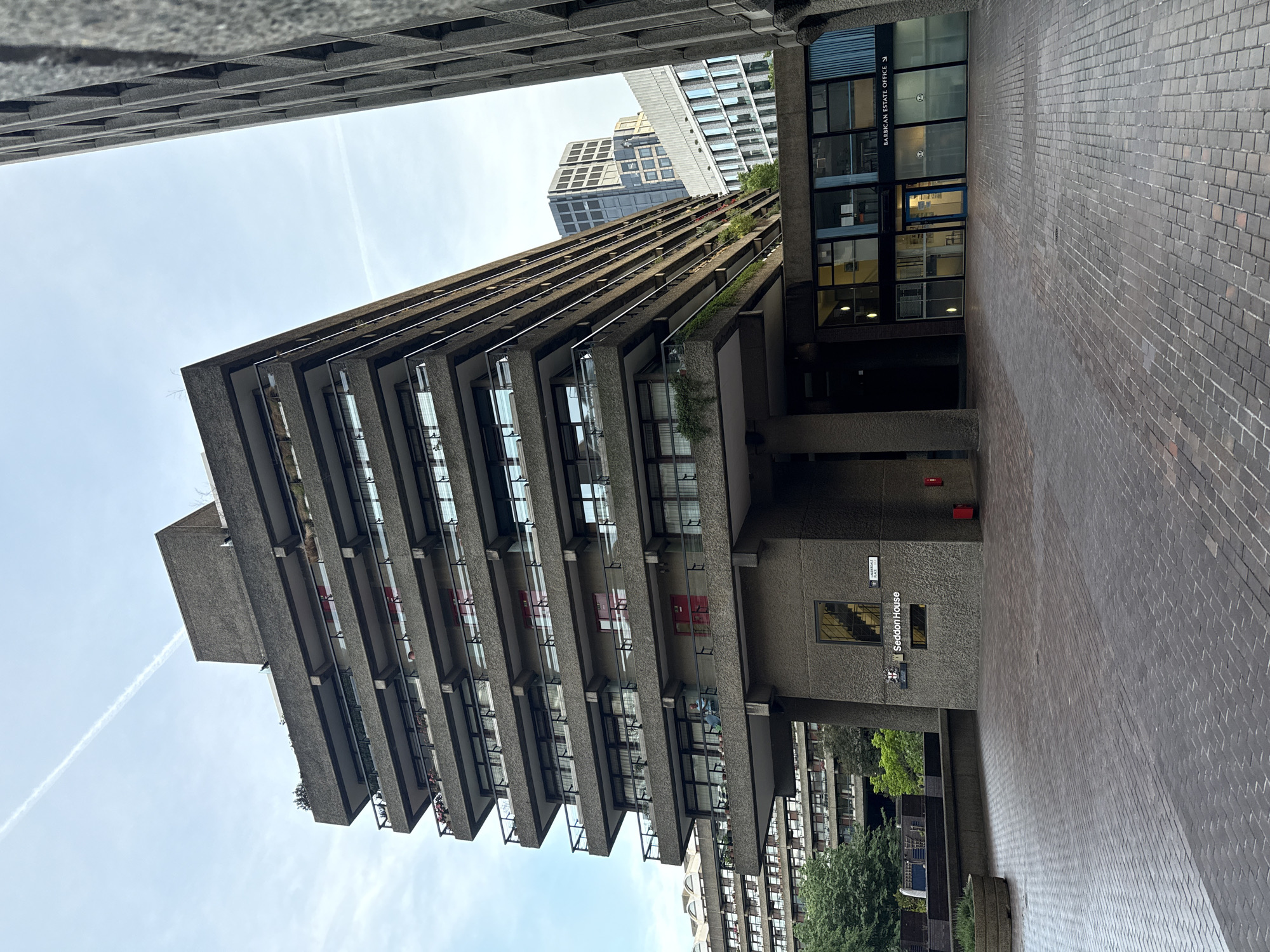 The image depicts the Brutalist architecture of the Barbican Estate in the City of London, United Kingdom. The frame is composed of a multi-story building with projecting concrete balconies and glass windows. In the foreground, there is a brick-paved walkway and a sign indicating "Seddon House". Another building with a glass storefront labelled "Barbican Estate Office" is adjacent to the building. A tall skyscraper is visible in the background, partially obscured by the multi-story building. The sky is visible above the buildings.