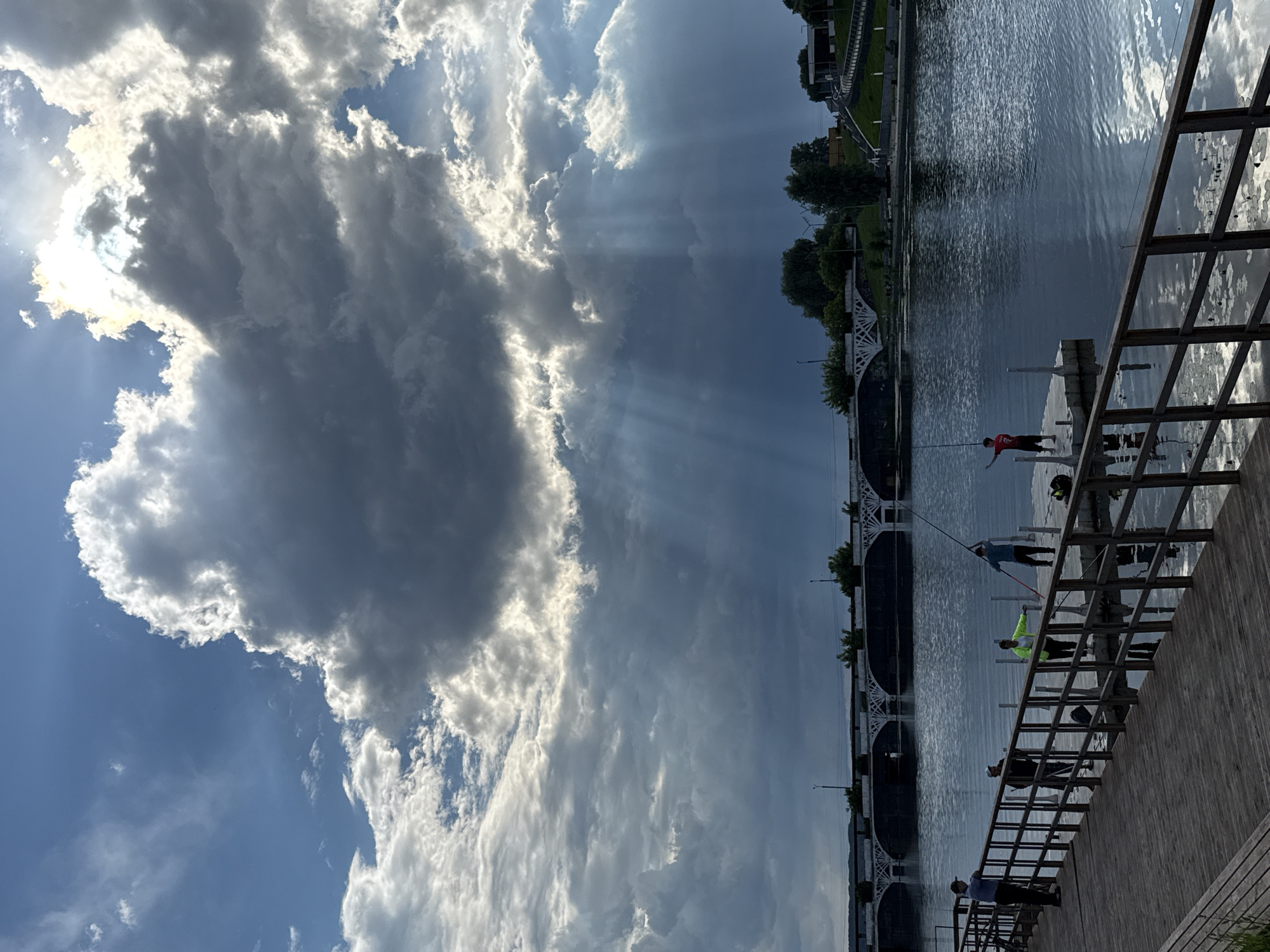 This is a photograph of the Votkinsk Reservoir in Votkinsk, Russia, on a cloudy day with dramatic sunbeams piercing through the clouds. The foreground features a wooden pier with a railing, leading out onto the water. Several people are visible on the pier and a smaller floating dock, most of whom appear to be fishing. One person on the floating dock is wearing a bright yellow jacket. Across the water, a bridge with multiple arches spans the reservoir, with trees and buildings visible on the far bank. The sun is partially obscured by large, dark clouds, creating a high-contrast scene with shafts of light illuminating parts of the water and the sky. The water is calm, reflecting the sky and the surrounding environment.