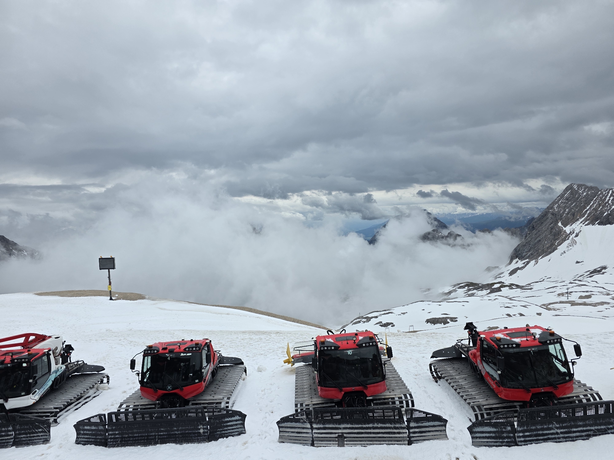 The image captures a high-altitude, snow-covered mountain landscape on the Zugspitze glacier plateau, located near Garmisch-Partenkirchen, Germany. In the foreground, four snowcats (pisten bullies) are parked in a row on a groomed snow surface. Three of these vehicles are predominantly red with black track systems and front blades, while the leftmost partially visible snowcat exhibits white and blue livery. Each snowcat features a cabin with multiple lights and a heavy-duty track system designed for snow traversal.
In the midground, a yellow pole with a dark, rectangular display screen is visible, positioned on the snow. The terrain consists of extensive snow fields with subtle undulations. The background reveals rugged, snow-capped mountain peaks partially obscured by dense, low-hanging grey clouds. Layers of clouds also fill the valleys and lower slopes of the distant mountains, creating a misty atmosphere. The sky above is entirely overcast with thick, grey clouds. No human figures are present in the scene.