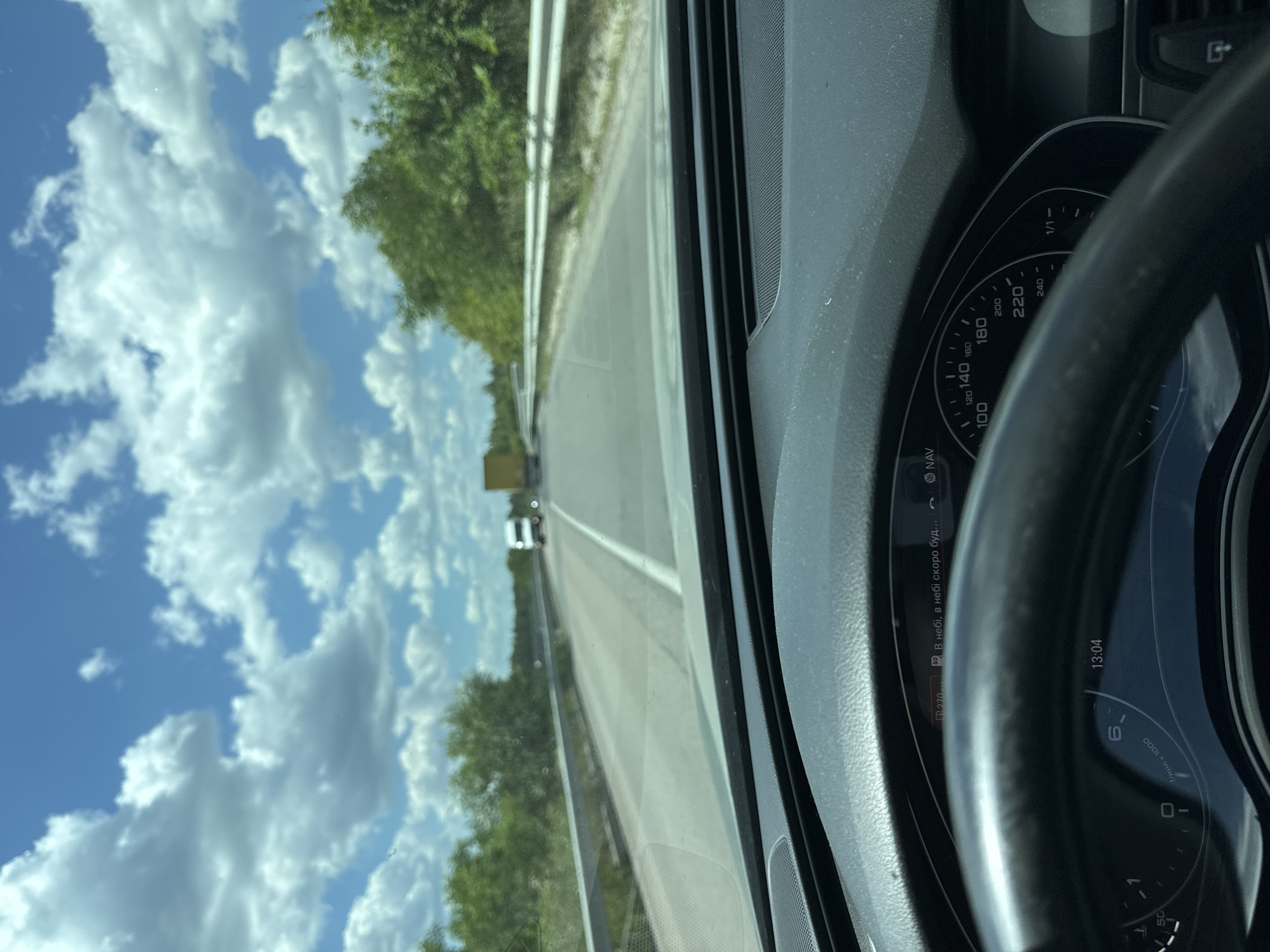 This media file is a first-person perspective shot from inside a car driving on a highway near Sarny, Ukraine. The view looks forward and to the left, capturing the road ahead, a guardrail, and a lush green tree line. The sky is a vibrant blue, dotted with fluffy white clouds, suggesting fair weather. In the distance, other vehicles are visible on the road, including what appears to be a truck.
The car's dashboard is prominently featured on the right side of the frame. It includes a speedometer with markings up to 240, and a tachometer. The time displayed on the dashboard is 13:04. Some text is visible on a small display screen, reading "• В небі, в небі скоро буд..." which translates to "• In the sky, it will soon be in the sky...". This could be a navigation prompt or a message from the car's system. The steering wheel is partially visible in the foreground. The overall impression is of a daytime drive through a scenic, possibly rural, landscape.