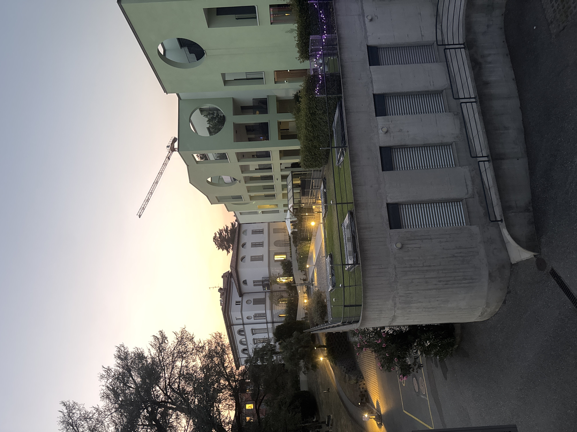 This image captures a twilight scene in Balerna, Switzerland, featuring modern apartment buildings with circular windows and a larger, older villa in the background. A construction crane is visible in the distance, suggesting ongoing development. The foreground shows a concrete structure with several windows covered by metallic blinds, leading to a walkway with a black metal railing. A street with yellow lines and circular manhole covers is visible to the left, illuminated by streetlights. Lush green vegetation and flowering bushes add a natural element to the urban landscape. The sky is a soft gradient of pale blue and orange, indicating the transition from day to night. No people are present in the scene.