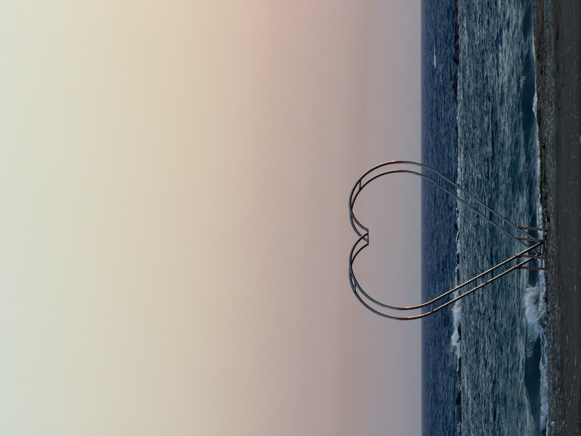 A large, metallic, heart-shaped sculpture stands on a pebble beach, facing the sea. The sculpture is positioned centrally in the lower half of the frame and is constructed from two parallel, curved metal tubes forming the heart shape, with supporting legs at the bottom. Beyond the beach, the deep blue sea is visible, with waves crashing against submerged rocks. The horizon line separates the sea from a gradient of soft pastel colors in the sky, transitioning from pale yellow at the top to a muted pink near the horizon, suggesting either sunrise or sunset. The scene is devoid of people. The location is identified as the coast of Meneou, Cyprus.