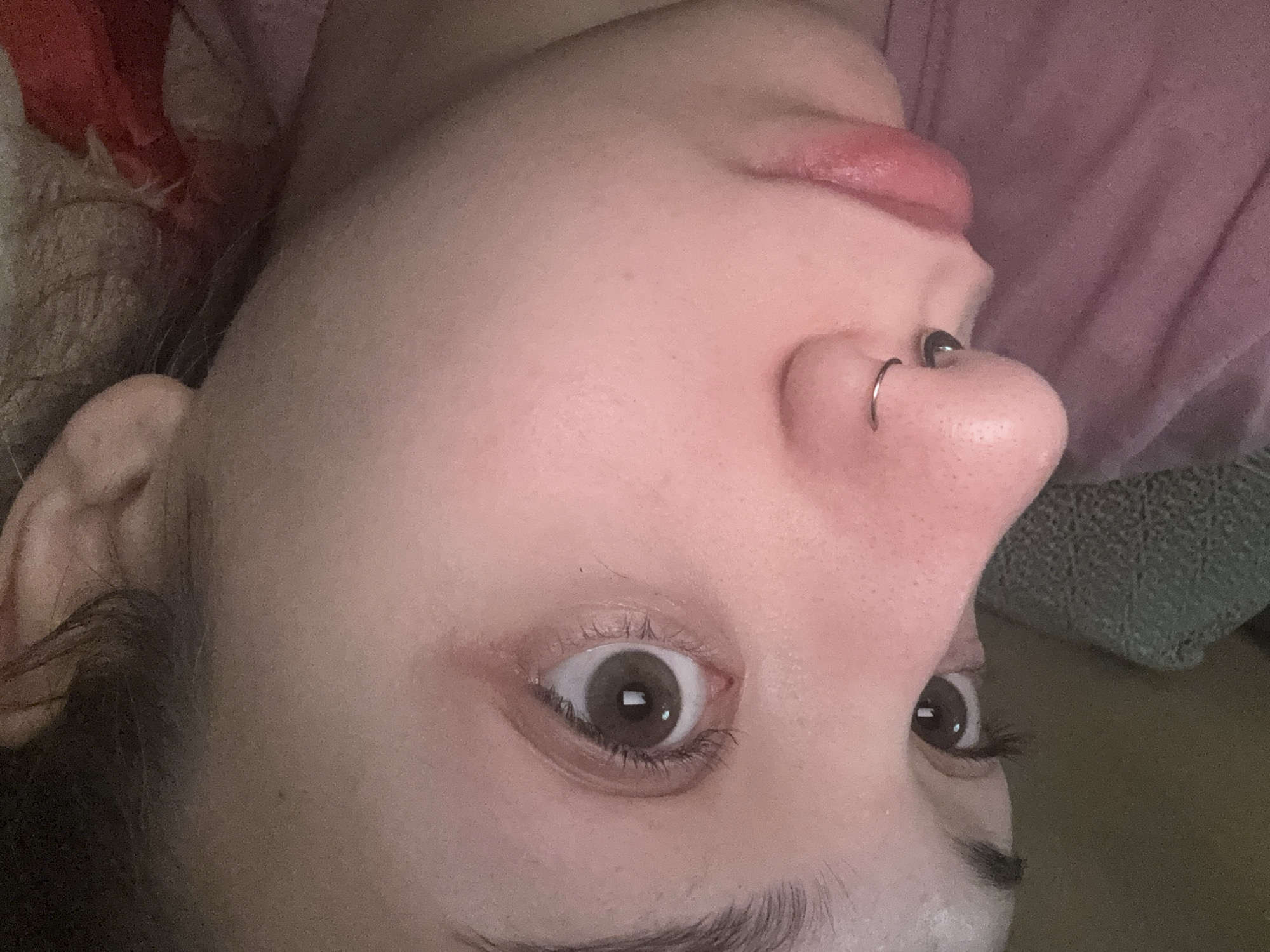 A close-up, angled, low-light photograph shows a person's face, focusing on the eye, nose, and lips. The person has brown eyes and a septum piercing. Their skin appears fair with some redness. The background is blurred and dark, with a hint of pink fabric visible. The location in Olaine, Latvia, is not determinable from the image.