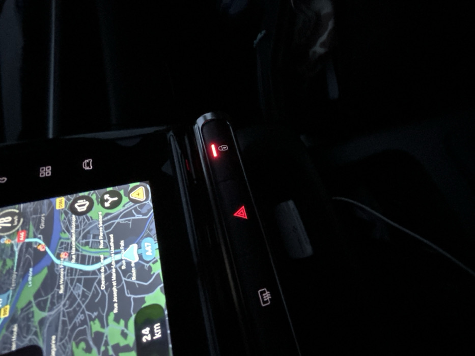 The image shows a car's dashboard and center console in low light. A navigation display with a map is visible, indicating a route with a current speed of 78 km/h and 24 km remaining. Controls to the right include a button with a red light indicating the car doors are locked, a hazard lights button (also with a red indicator), and a button for window defrost. The surrounding area appears to be dark and possibly cluttered.