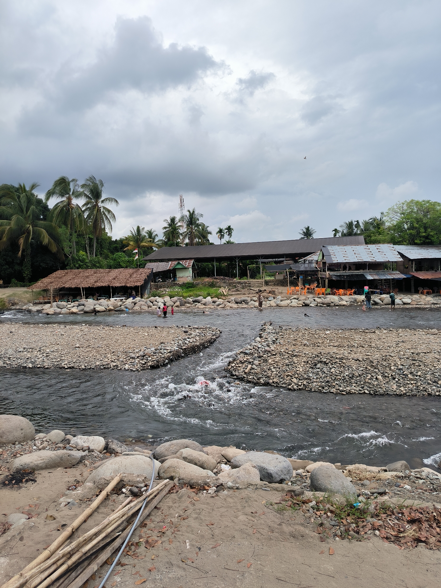 The image depicts a lively river scene in Meurah, Indonesia, under an overcast sky filled with soft grey clouds. A broad, shallow river with a rocky bed dominates the midground, its dark waters showing areas of white rapids and gentle flow. Numerous large, smooth rocks are scattered across the riverbed and form natural and possibly man-made barriers, creating narrower channels for the water.
On the left bank, nestled among abundant palm trees and tropical vegetation, stands a rustic open-air structure with a distinctive thatched roof, serving as a shaded eating or resting area with tables and chairs visible. Several individuals are gathered here, and others are wading in the shallow river water nearby.
Further along the right bank, a series of more substantial buildings with corrugated iron roofs line the riverside. These structures also feature outdoor seating arrangements with numerous orange and other colored plastic tables and chairs placed on rocky platforms overlooking the water, suggesting they are riverside cafes or eateries. A few people, including children, are seen standing and playing in the shallow parts of the river adjacent to these establishments.
In the background, a dense cluster of lush green trees, including many palm trees, forms a verdant backdrop. A tall telecommunications tower is visible rising above the trees on the right. A small Indonesian flag (red and white) can be seen flying on a pole near one of the buildings, indicating the country.
The immediate foreground features a sandy bank with scattered dry leaves, a pile of bamboo poles, and a grey pipe, suggesting some local material or infrastructure. The overall scene conveys a relaxed, recreational atmosphere, with people enjoying the river in a natural, somewhat rustic setting. No visible text is legible in the image.