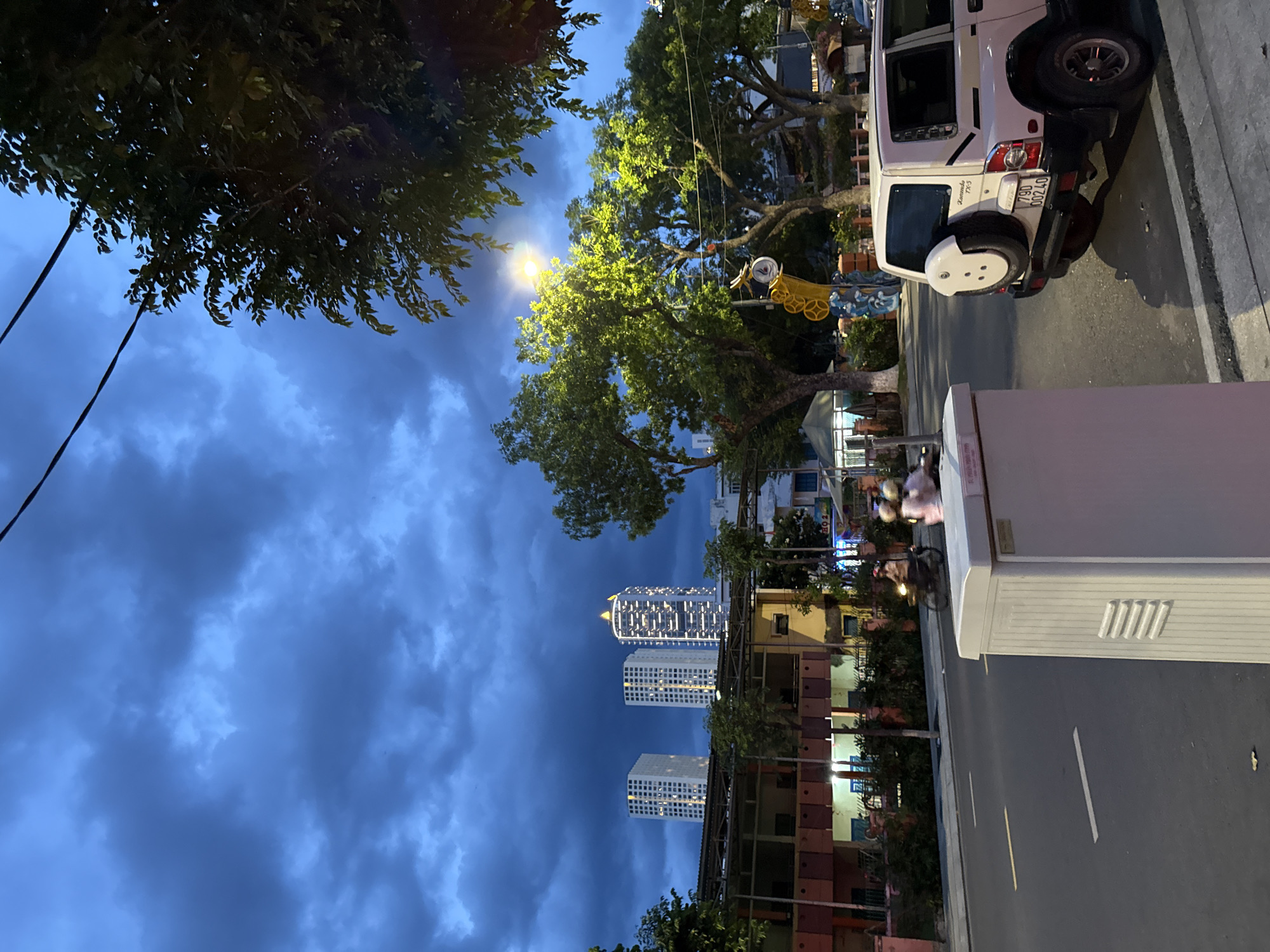 The image depicts a street scene at dusk in Nha Trang, Vietnam. The sky is filled with dark, dramatic clouds, suggesting an approaching storm. In the background, three tall, modern buildings are illuminated, with one in the center appearing particularly brightly lit. The street in the foreground is asphalt with white lane markings. On the right side of the street, a white SUV is parked, facing away from the camera. A light pole with a bright yellow glow illuminates some of the trees. On the left side of the street, a large white utility box is prominent in the foreground. Further down the street, a motorcycle with two riders is visible, their faces obscured by helmets. The overall atmosphere is moody and somewhat urban.