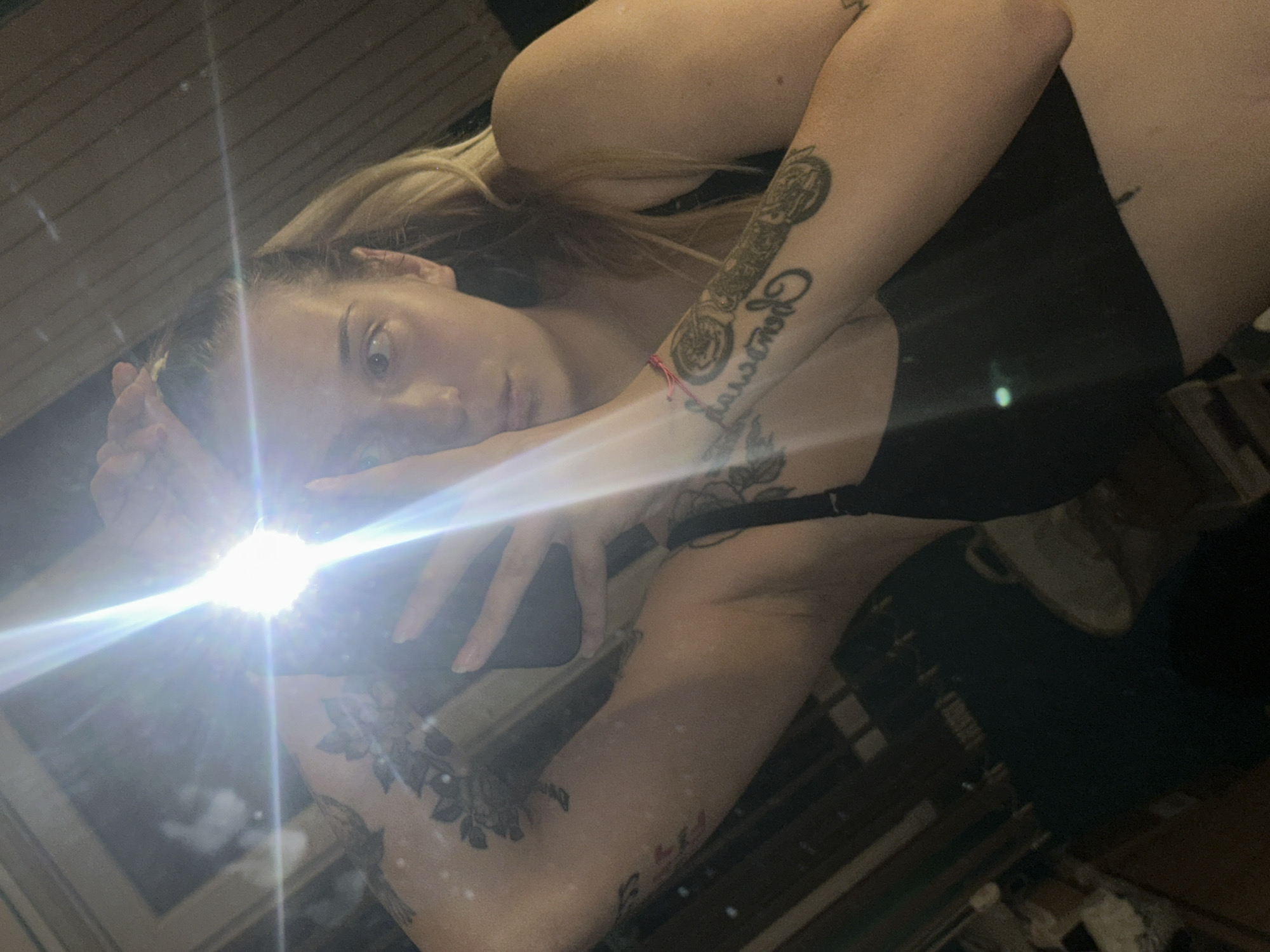 The image is a selfie taken in a dimly lit room, possibly a bedroom, given the presence of what appears to be a wooden-paneled wall and furniture in the background. A strong light source, likely a camera flash or a very bright lamp, creates significant lens flare and washes out parts of the image. The subject is a young woman with blonde hair tied back in a ponytail. She is wearing a black tank top or bra. Her arms are raised, with one hand near her face and the other holding a phone, suggesting she is taking the picture. She has several tattoos visible on her arms, including floral designs and what appears to be an illustration of a motorcycle. The woman is looking towards the camera with a neutral expression. There are no clear indicators of the location being in Plovdiv, Bulgaria, as the image lacks any recognizable landmarks or cultural elements specific to that city. The overall scene is personal and intimate, typical of a selfie taken in a private space.