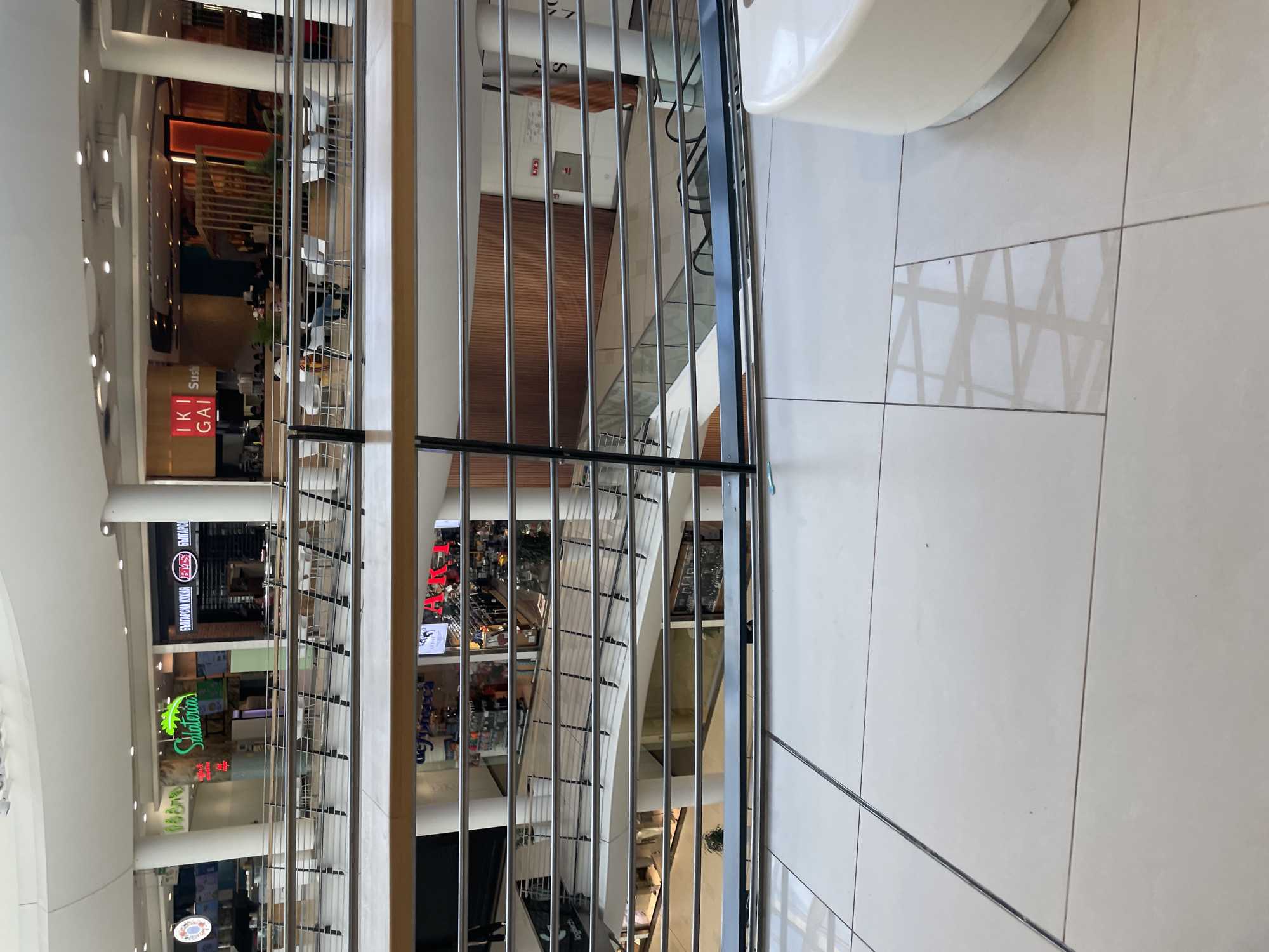 The image depicts an interior view of a shopping mall in Sofia, Bulgaria, taken from an elevated position, likely a balcony or upper floor. The foreground features a metal railing with vertical bars. Through the railing, multiple levels of the mall are visible, with various storefronts including "IKI GAI Sushi," "Salateria," and "Българска Кухня". The ground floor is populated with shops and possibly customers, although individual people are not clearly discernible. The mall interior features white tile flooring, white pillars and ceilings. The light is bright, suggesting natural light or strong artificial illumination. Escalators are visible between the floors. The overall impression is a modern, multi-level shopping center.