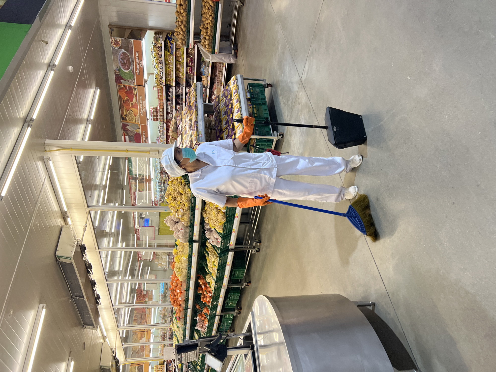The image depicts an interior scene of a supermarket in Baan Bo Phut, Thailand. A person wearing a white uniform, a white cap, a light blue mask, and orange gloves is sweeping the concrete floor with a blue-bristled broom. They are holding a black dustpan in their other hand. The supermarket aisle contains shelves stocked with various produce, including tomatoes, onions, potatoes, and other unidentifiable vegetables and fruits. In the background, more shelves are visible, displaying packaged goods. The lighting in the supermarket appears to be fluorescent, with visible light fixtures on the ceiling. The overall environment suggests a commercial retail space focused on selling groceries.