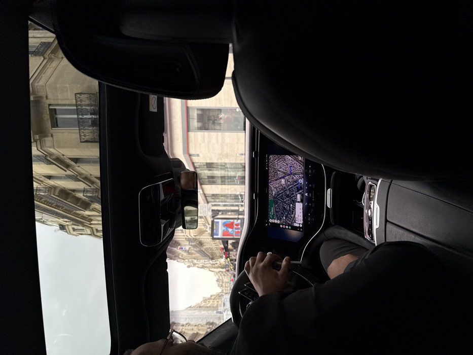 The image depicts the interior of a vehicle from a rear passenger's perspective, looking forward through the windshield and partially through the sunroof. A male driver, wearing glasses, a dark jacket, and dark shorts, is seated on the left, with his right hand on the steering wheel and his left hand resting near the central console's gear selector. The car's infotainment screen displays a navigation map identifying the current location in the Opéra district of Paris, France, specifically showing street names such as Rue des Capucines and Boulevard des Capucines.
The exterior view through the windows reveals a classic Parisian street lined with multi-story Haussmannian buildings, characterized by light-colored stone facades and black wrought iron balconies. An overcast sky is visible overhead. On the right side of the street, a distinctive red and blue Morris column stands. The vehicle's interior features dark upholstery, a prominent central display screen, and a visible control panel on the center console.