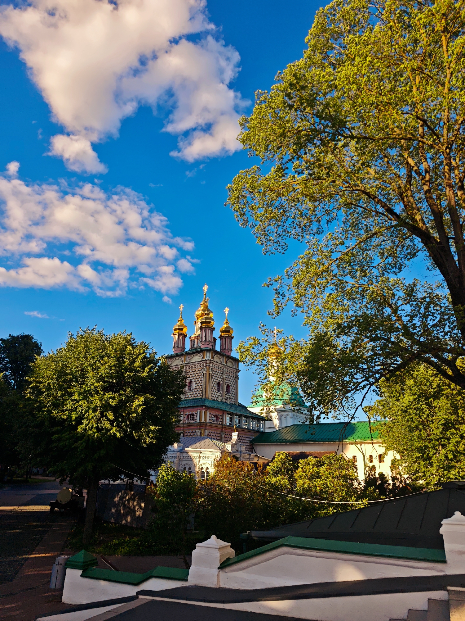 The image shows a vibrant, sun-drenched scene of a Russian Orthodox church with golden domes, peeking through lush green trees. The sky is a brilliant blue, dotted with fluffy white clouds, suggesting a pleasant, clear day. The sun's warm light casts long shadows and illuminates the leaves of a large tree in the foreground, highlighting its texture and vibrant green color.
The church itself is ornate, with intricate architectural details and a mix of pink, white, and green facades. Several golden domes rise majestically against the sky, topped with crosses, characteristic of Orthodox Christian churches. The surrounding greenery includes well-maintained trees and bushes, hinting at a park-like or monastic setting. In the lower part of the image, white decorative fencing and a dark roofline are visible, suggesting a pathway or a viewpoint. There are no people clearly visible in the image, and no text is present. The overall mood is serene and picturesque, evoking a sense of peace and spiritual grandeur. The lighting suggests it might be late afternoon or early morning, given the long shadows and the warm quality of the light. The location appears to be a place of historical and religious significance, possibly a monastery or a significant church complex.
