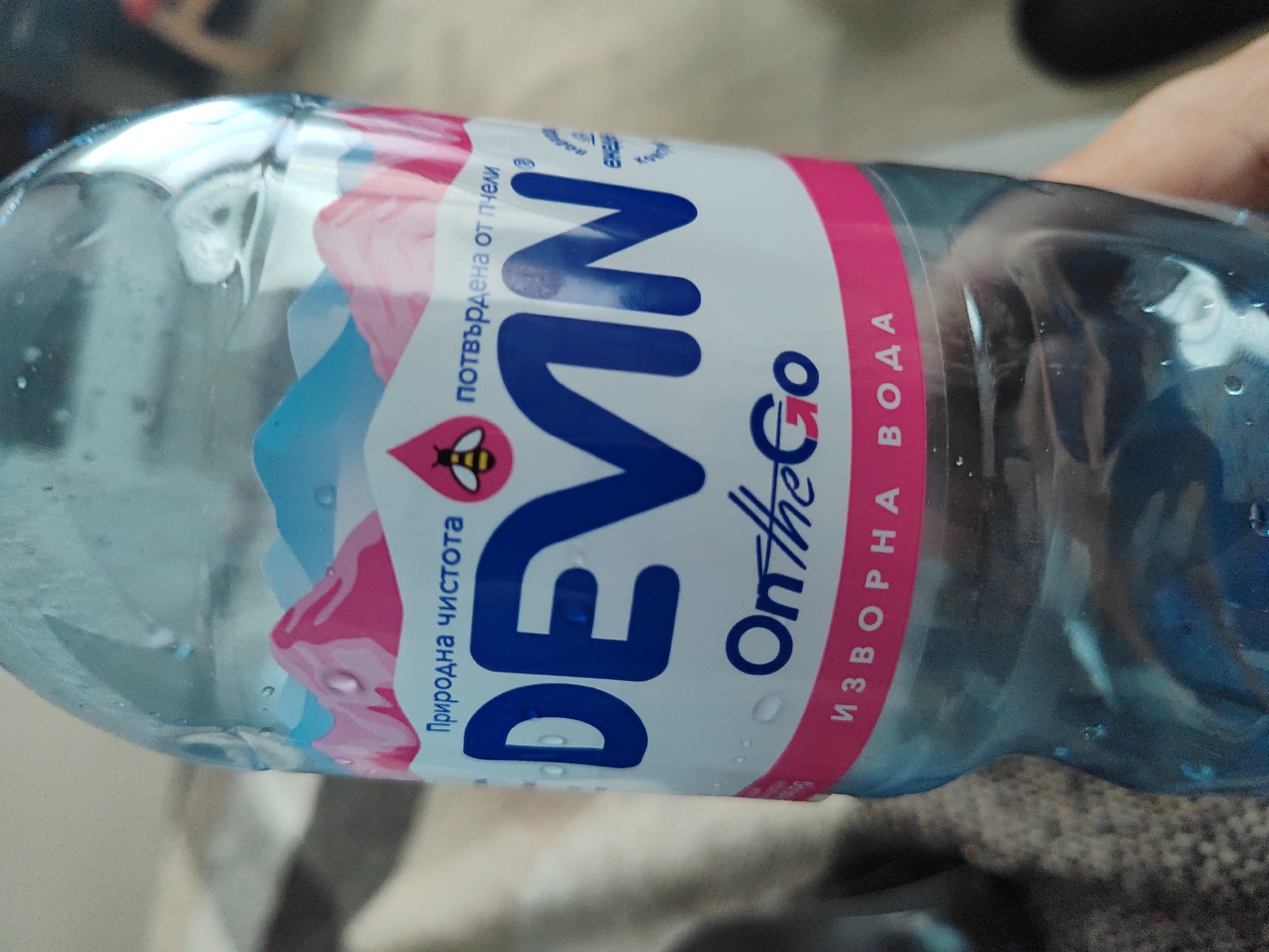 A close-up shot shows a plastic water bottle labeled "Devin On the Go". The label includes Bulgarian text "Природна Чистота" (Natural Purity) and "Изворна Вода" (Spring Water). The brand's logo features a bee. The location, Kaspichan, Bulgaria, is not discernable from the image. No people are fully visible, though a hand is partially present in the background. The environment includes a neutral-toned surface underneath the bottle.