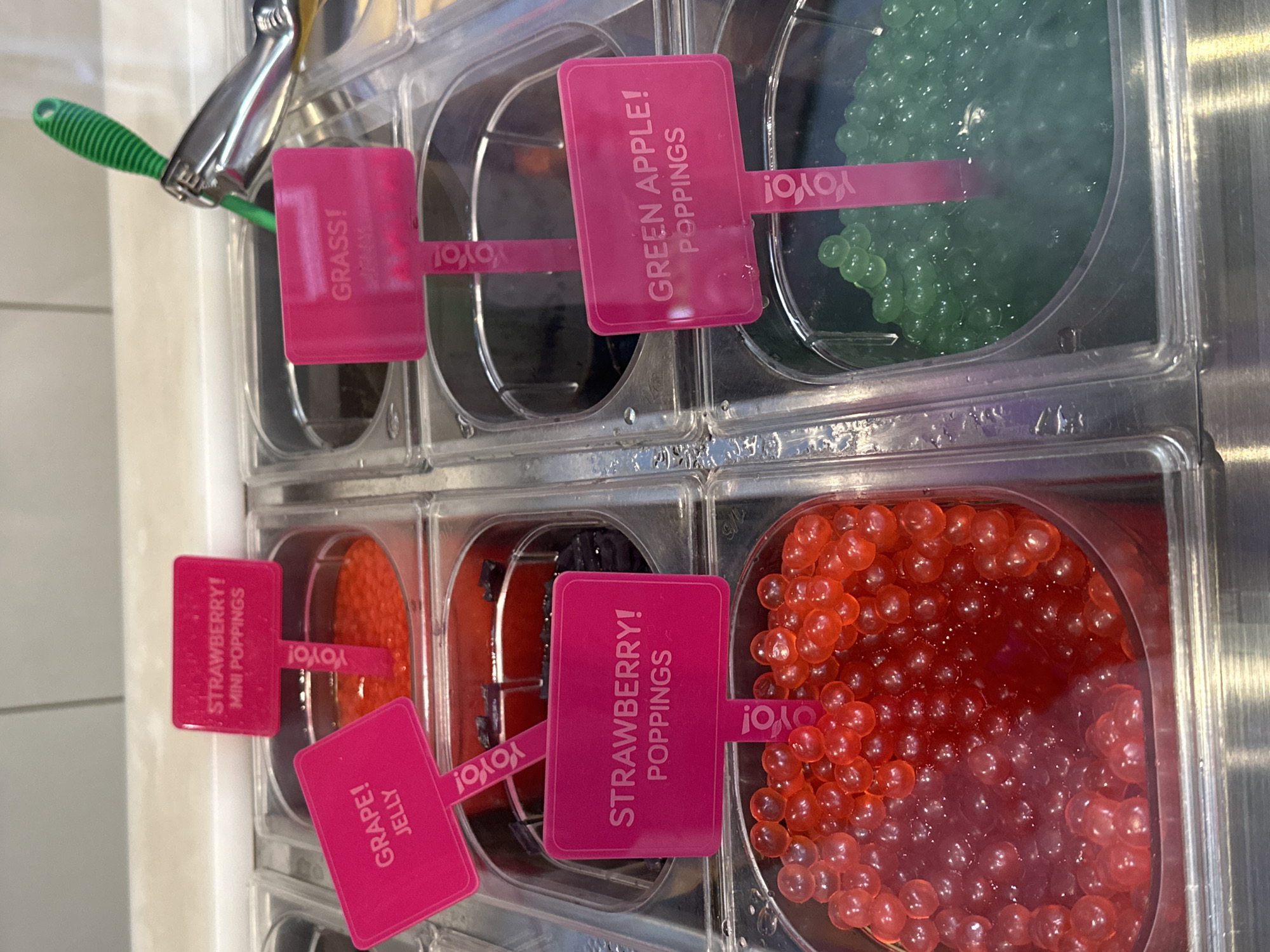 The image shows a close-up of several translucent, brightly colored popping boba toppings in clear plastic containers. Pink signs with white text are placed in each container, indicating the flavors. From left to right, the visible flavors are "GRAPE! JELLY", "STRAWBERRY! MINI POPPINGS", "GRASS!", and "GREEN APPLE! POPPINGS". Some of the containers appear to hold liquid or syrup beneath the boba. A metal scoop with a green handle is visible in the top right portion of the image, resting on the edge of a container. The scene suggests a self-serve frozen yogurt or dessert shop. The environment appears to be indoors, with a tiled or laminate countertop in the background. There are no people or distinct architectural features visible that would allow for an identification of the city or country.