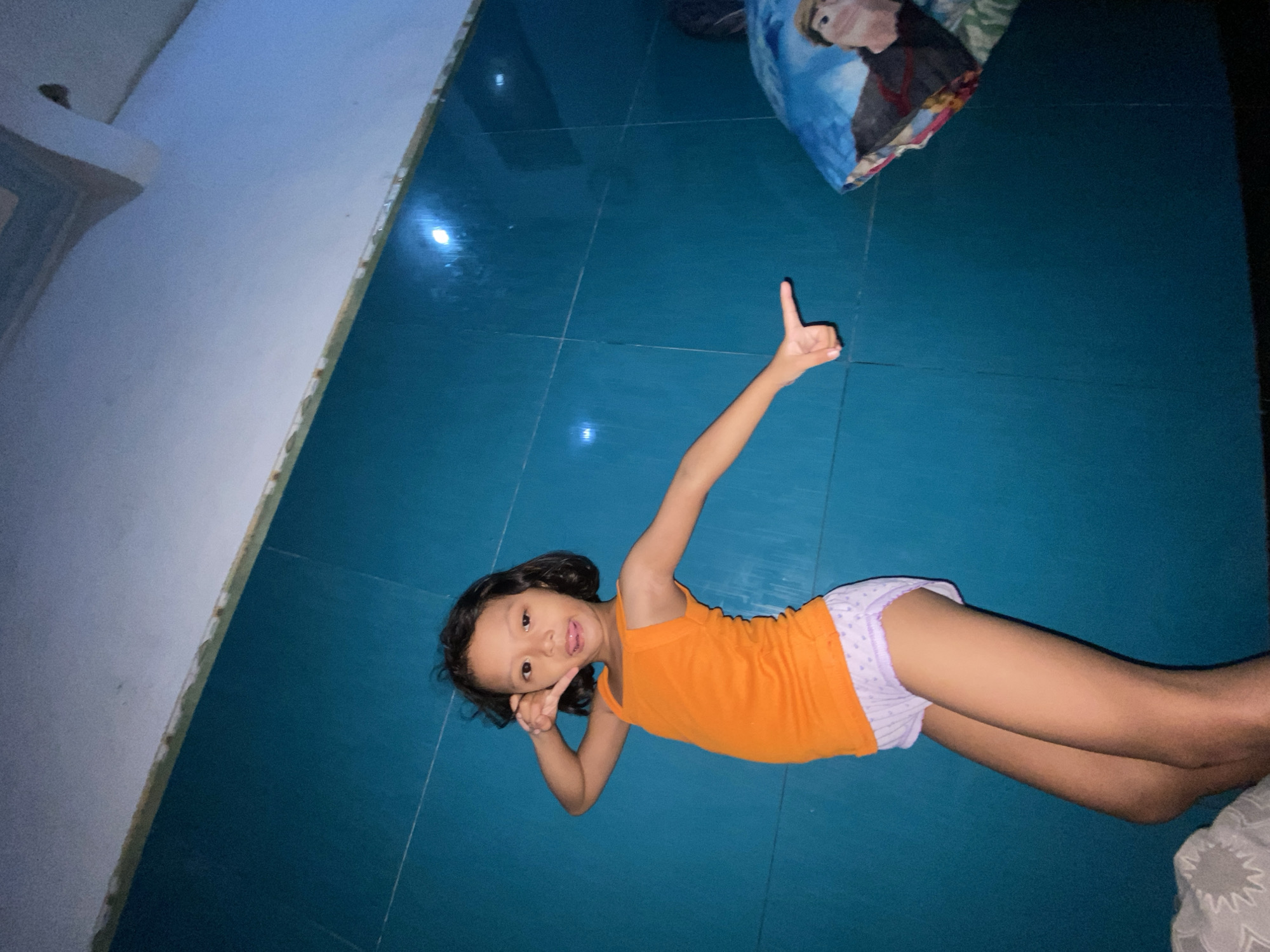 A young female child lies on a blue-tiled floor, with her head near a white wall. She is wearing an orange tank top and lilac polka-dot shorts. The child is making the "number one" gesture with one hand raised in the air, and making a "peace" sign with the other hand near her face, and sticking her tongue out. There is a pillow with a cartoon character printed on it in the top right corner of the image, and part of another pillow is in the bottom right corner. Based on the lighting and surrounding details, the location appears to be indoors, possibly a residential space in Gelam, Indonesia.