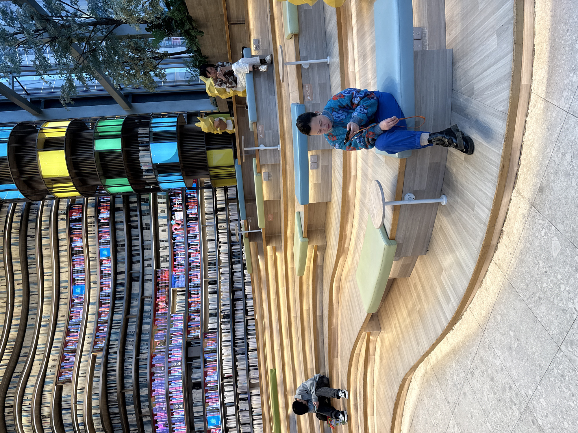 The image depicts the interior of a multi-story modern building, identified as a cultural or library space, located in Liangjiang, China. The scene is dominated by a massive, curved wall of bookshelves on the left, extending vertically for multiple floors. These shelves feature integrated strip lighting and display a mix of physical books and illuminated panels simulating colorful book spines. Some shelves also incorporate digital screens displaying content.
In the foreground and mid-ground, a tiered, undulating seating area made of light-colored wood occupies the central and lower right sections. This terraced structure includes various benches with pale blue and light green cushions. Small round tables with white bases are interspersed among the seating platforms, which also feature integrated strip lighting along their edges. Power outlets are visible on some of the seating risers. The floor in the immediate foreground is covered with light gray speckled tiles.
Three individuals are present, each engaged with a smartphone and not interacting with each other:
1. **Foreground, center-left**: A female subject with short dark hair sits on a light blue bench. She wears a multi-colored patterned sweater (blue, orange, red, white) and blue pants, with heavy black boots. White earbuds are visible in her ears as she looks down at her smartphone.
2. **Mid-ground, left**: A male subject sits on a wooden step, looking down at his smartphone. He wears a grey hooded sweatshirt, dark pants, and black and white sneakers. A small colorful bag rests on the step beside him.
3. **Background, right**: A female subject sits on a light blue bench on a higher tier. She wears a brown and white plaid shirt and white pants. She is also looking down at her smartphone. Behind her, near a wall and lush green plants, two large, yellow, polygonal animal-like figures are positioned.
To the right of the main seating area and extending vertically is a prominent cylindrical, multi-tiered structure, possibly a core or elevator shaft. This structure is adorned with rectangular panels that emit yellow, green, and blue light. Adjacent to this cylinder on the far right, large windows reveal an outdoor area with trees and additional foliage.