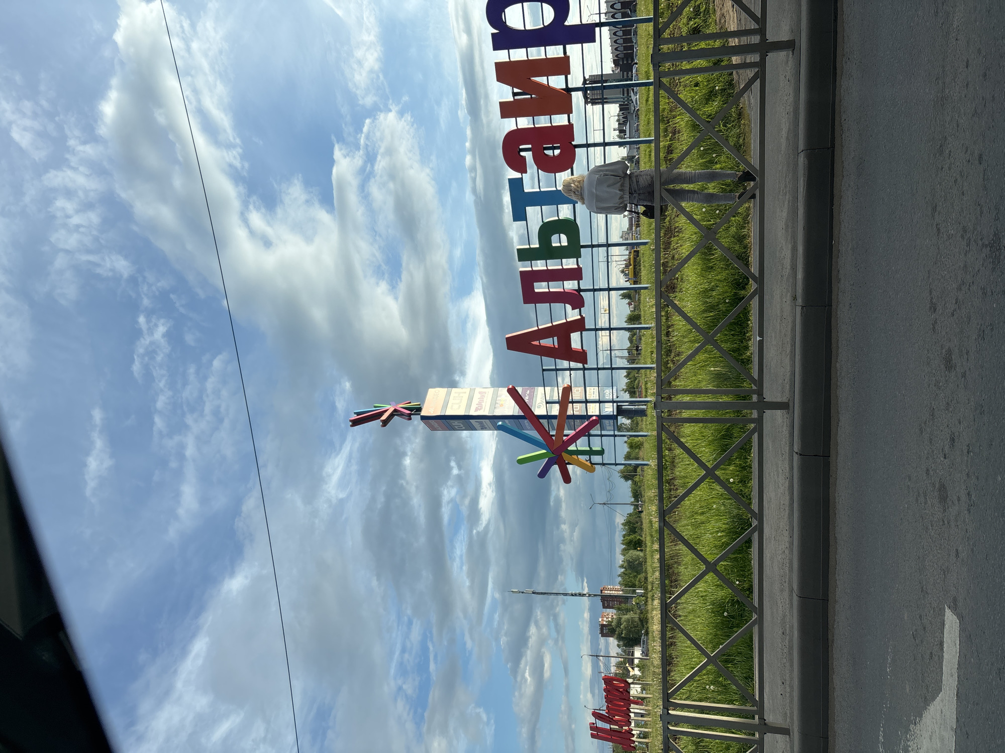 The image depicts a street scene in Yaroslavl, Russia, featuring a large, colorful sign with the name of the city in Russian Cyrillic letters. The sign is positioned near a metal fence bordering a grassy area. A person is standing by the fence, seemingly observing the scene. In the background, buildings and greenery are visible under a cloudy sky. An advertisement tower and a street lamp pole are also noticeable in the background. A roadway occupies the foreground.