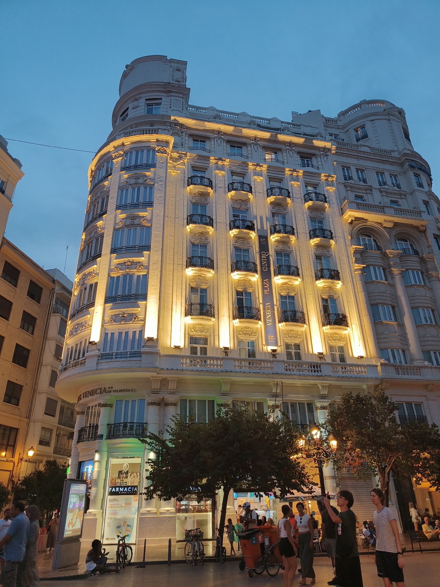 The image depicts the Hotel Venezia de Valencia S.L. located in the city of Valencia, Spain. It is a nighttime shot of a grand, cream-colored building with ornate architectural details and multiple balconies with black wrought-iron railings. The building is illuminated by warm yellow lights, highlighting its facade. In the foreground, there are trees, streetlights, and a few bicycles. People are visible on the sidewalk, some walking and others standing. A pharmacy, identified as "FARMACIA," is situated at the ground level of the building, with its windows lit. A Taco Bell advertisement is also visible. The sky is a dark, dusky blue, indicating it is either dawn or dusk.