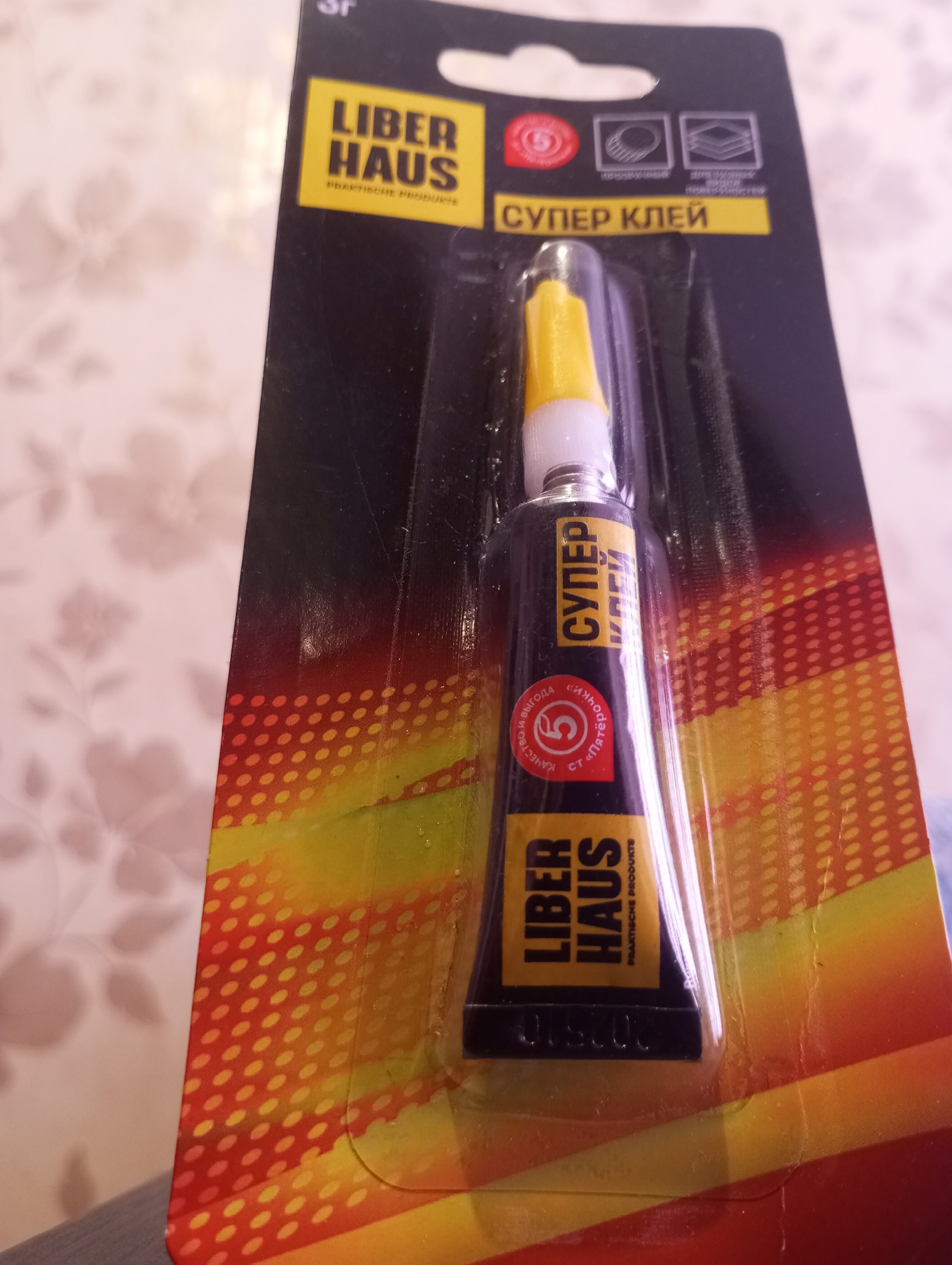 The image shows a retail package of "Super Glue" by the brand "LIBER HAUS PRAKTISCHE PRODUKTE". The packaging is primarily black with yellow and orange accents, featuring graphics and text in Russian. A small tube of glue with a yellow applicator tip is visible within the plastic packaging. The packaging is hanging on a display, suggesting it is for sale. The background appears to be a lightly patterned wallpaper. There is no discernible information or imagery in the photograph that would indicate the location in the city of Kirzhach, Russia.