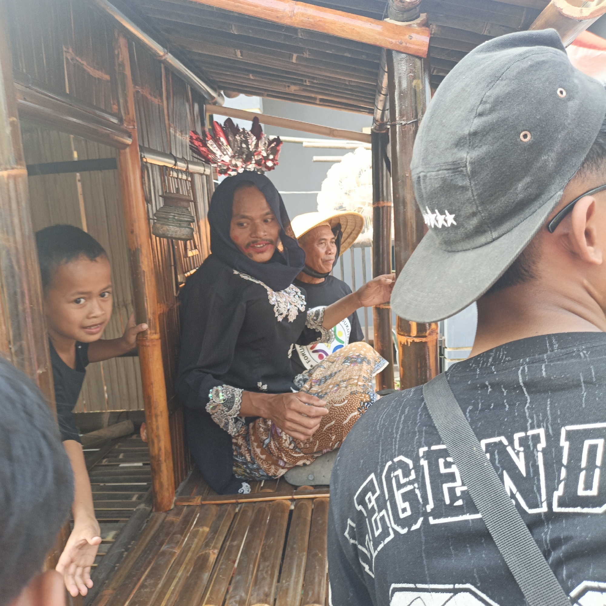 The image depicts an outdoor scene within a bamboo structure in Tanahbaya, Indonesia. In the center, a person wearing a black hijab and traditional patterned skirt is seated, with red and white headwear adorned with feathers above them. To their right, another person wearing a conical hat and a black shirt is visible. A young boy is seen looking towards the viewer from the left side of the frame. In the foreground, the back of a person wearing a black cap and a t-shirt with white lettering is partially visible. The structure itself is constructed from bamboo poles and slats, with a thatched or woven roof. The overall impression is of a cultural or ceremonial setting.