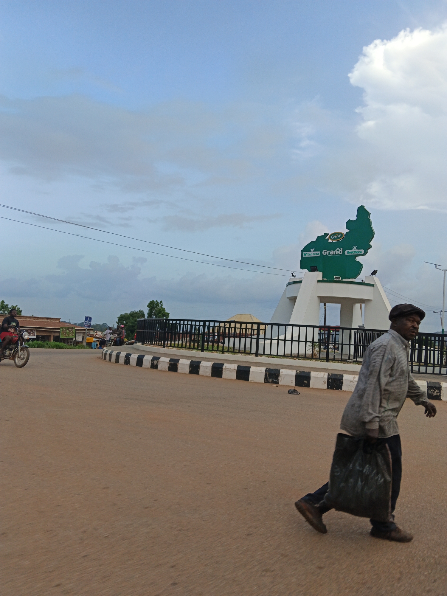 In Kuru Dungus, Nigeria, a man wearing a cap and a checkered long-sleeved shirt walks from right to left across a paved road, carrying a black plastic bag in his right hand. He wears dark pants and brown shoes. In the background, a green map-shaped monument with the word "Grand" and "Vital Feed" inscribed on it is visible. The monument is white at the base and has a black fence around it. A few motorcycles are also present. The sky is cloudy, with some patches of blue. The overall scene depicts a public area with a landmark and pedestrian activity.