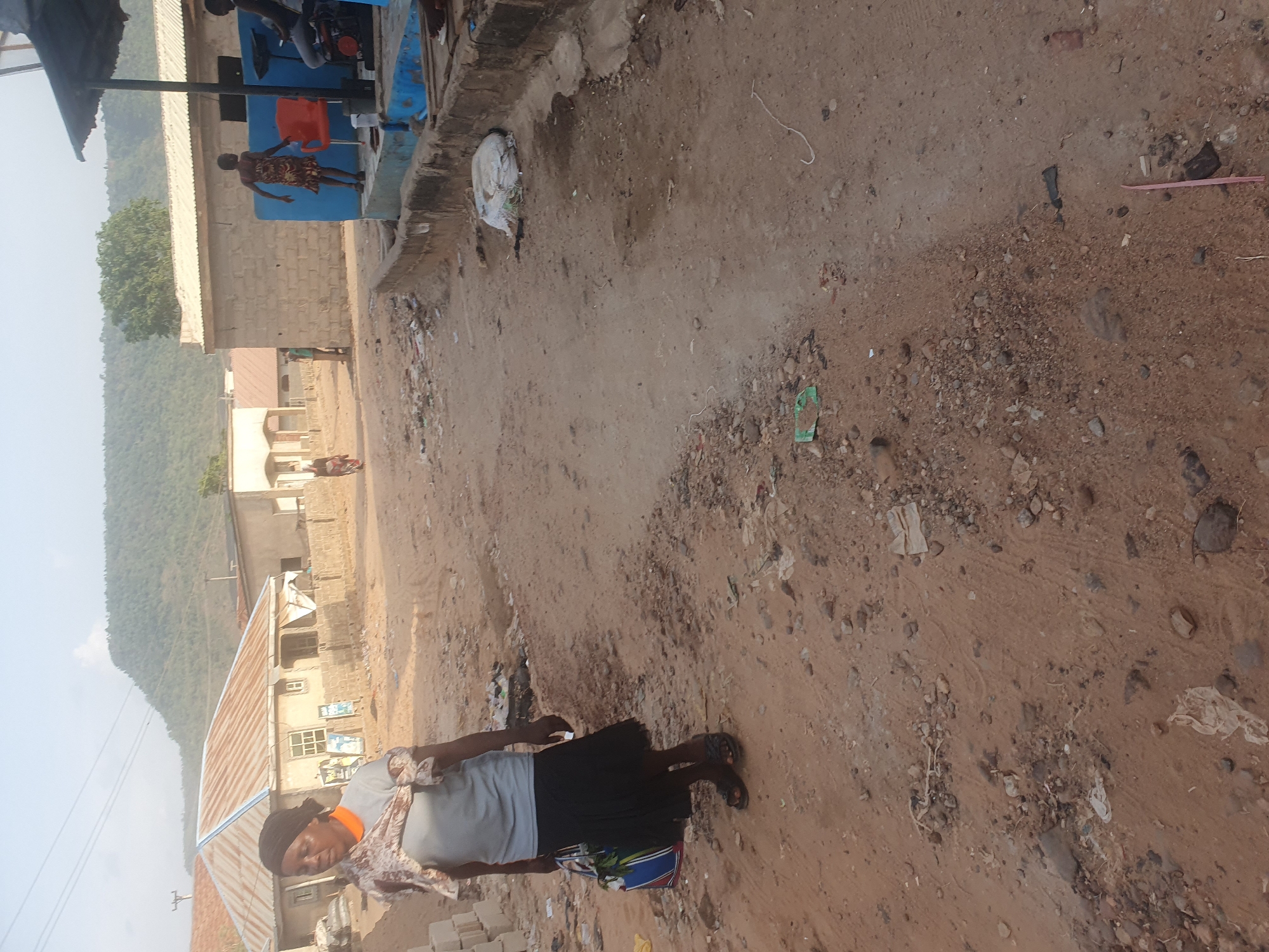 A woman wearing a gray shirt and a black skirt is standing on a dirt road in Lokoja, Nigeria. The road is lined with buildings and a mountain can be seen in the background. There is litter on the ground. Several other people are visible in the distance. A motorcycle is parked near a blue structure in the upper portion of the image.