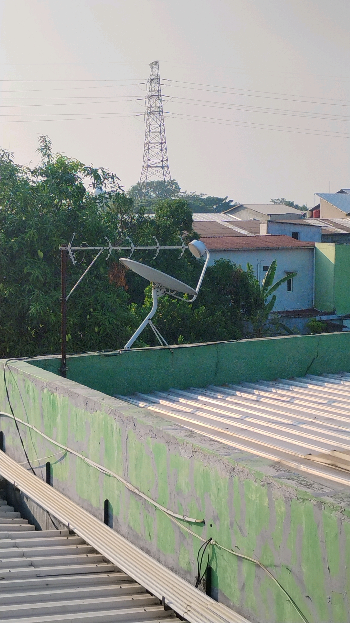 The image depicts a residential rooftop scene in Gumpang, Indonesia, on a clear, bright day. In the foreground, a green-painted concrete wall with visible cracks and peeling paint runs horizontally across the frame. Several corrugated metal roofs are visible on either side of the wall.
Dominating the mid-ground is a satellite dish mounted on a white stand, positioned on the rooftop. To its left, a metallic antenna with multiple horizontal rods is attached to a vertical pole. Behind the satellite dish and antenna, dense green foliage from trees obscures much of the background.
Further in the background, a cluster of houses with various roof colors, including red and light blue, are visible. Beyond the houses, a tall, lattice-style electricity pylon stands prominently against the pale sky, with power lines extending from it. The overall impression is one of a typical urban or semi-urban Indonesian environment, with a mix of domestic technology and essential infrastructure.
