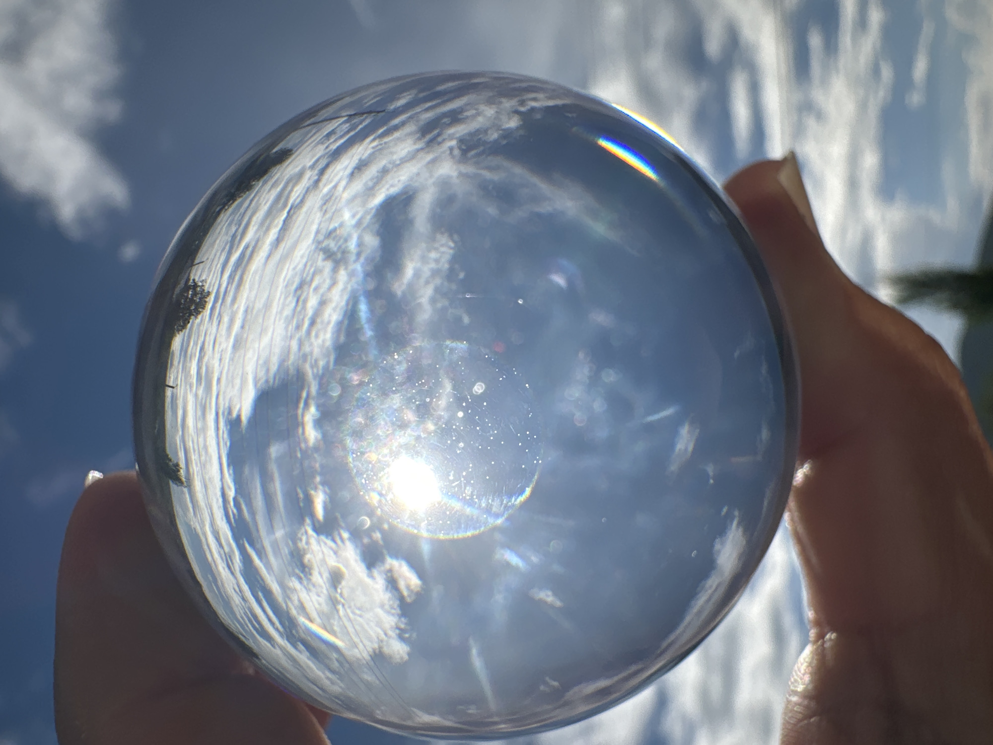 A light-skinned hand holds a clear, spherical crystal ball against a bright outdoor backdrop of a blue sky with scattered white cumulus clouds. The sphere's surface reflects and refracts the ambient light and environment, presenting an inverted and compressed view of the sky and clouds within its interior. A powerful sunburst or specular highlight emanates from the upper-central region of the sphere, causing significant glare, spherical aberrations, and visible small, circular bokeh lights and faint multicolored lens flares throughout the sphere's volume. Portions of the hand's fingers are visible, gripping the sphere from its upper and lower extremities. Outside the sphere, the upper background displays an expanse of deep blue sky with distinct white clouds. The lower background features additional sky and a blurred, dark green element, likely vegetation or distant terrain. A prominent rainbow-colored prismatic effect is visible along the lower right curvature of the sphere. The scene is brightly illuminated by direct sunlight. The location is Unknown, Unknown.