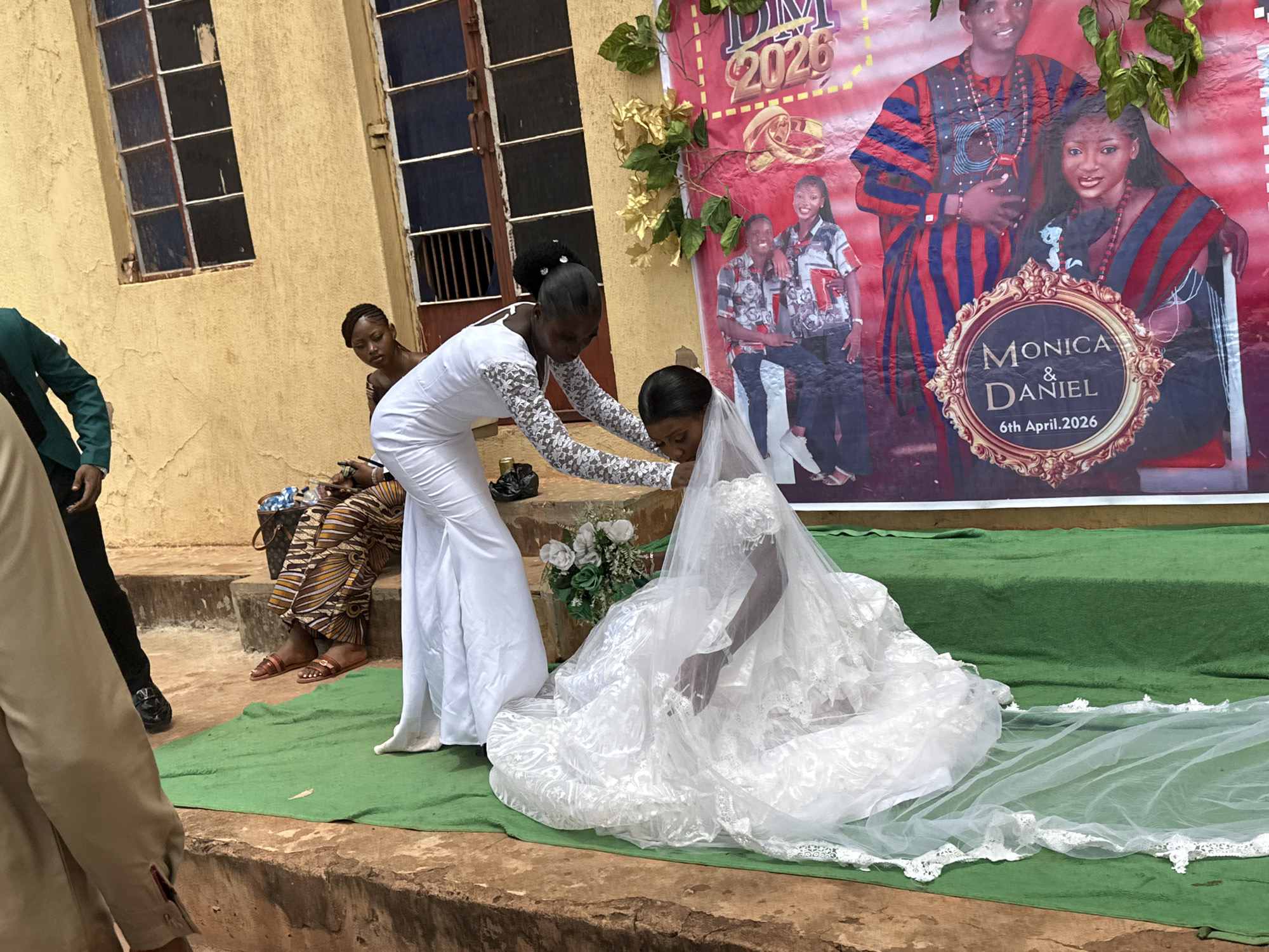 The image captures a wedding event in an unidentified urban location, with a focus on the bride and her attendant. The bride, dressed in a voluminous white wedding gown with lace detailing and a long veil, is kneeling on a green carpet. Her attendant, also in a white dress with lace sleeves, is adjusting the bride's veil. Behind them, a backdrop banner featuring the couple, labeled "Monica & Daniel 6th April, 2026", suggests the event is a wedding reception or photo opportunity. To the left, a woman is seated, observing the scene, and sections of men in suits are also visible. The environment includes a yellow building exterior with barred windows and what appears to be an outdoor stage or platform area.