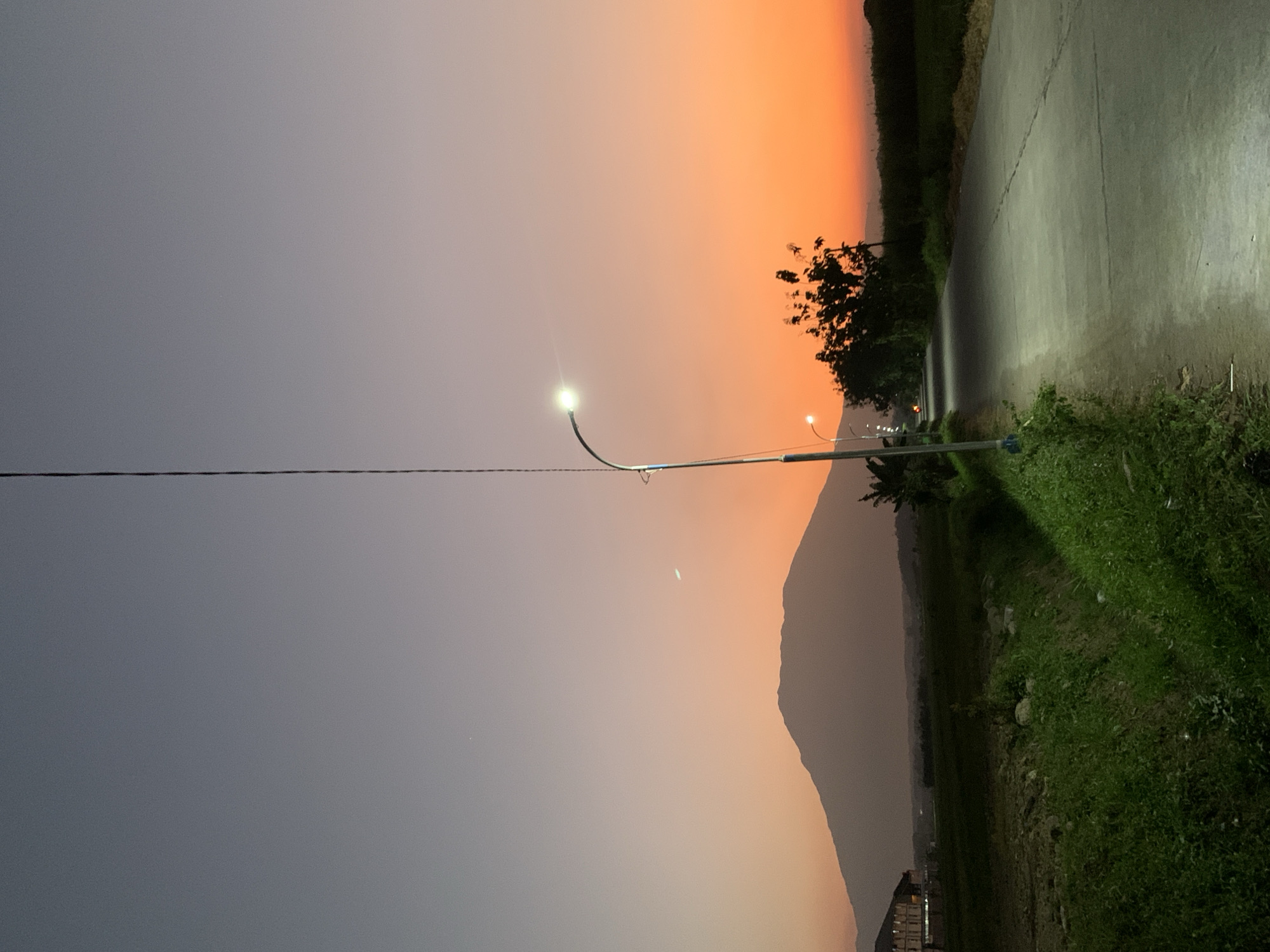 This detailed image captures a rural or semi-rural landscape at dusk in Jungke, Indonesia. The sky displays a striking gradient, transitioning from a dark grey-blue at the zenith, through soft lavender and pink mid-tones, to a vivid orange-red hue near the horizon. A faint, crescent moon is visible in the upper-left quadrant, with a small, bright star-like object positioned above and slightly to its left.
In the background, a large, dark mountain dominates the skyline, its silhouette sharply defined against the colorful sky. To the far left, a multi-story building with illuminated windows is partially visible at the base of the mountain.
The midground features a paved road, likely asphalt or concrete, extending from the foreground and curving slightly to the left as it recedes into the distance. The road surface exhibits some cracks. Several streetlights illuminate the path; a prominent streetlight on a tall pole is positioned on the left side of the road in the foreground, casting a bright, white light downwards. Another illuminated streetlight is visible further along the road, and the glow from additional lights can be seen.
The immediate foreground on the left consists of a grassy verge with lush green foliage, bordering a shallow ditch or embankment. Along the road, trees with dark, dense foliage are silhouetted against the bright horizon. No individuals or animals are present in the visible scene. A thin, dark, vertical line, possibly an antenna or utility wire, extends from the top edge of the image, running parallel to the foreground streetlight pole.