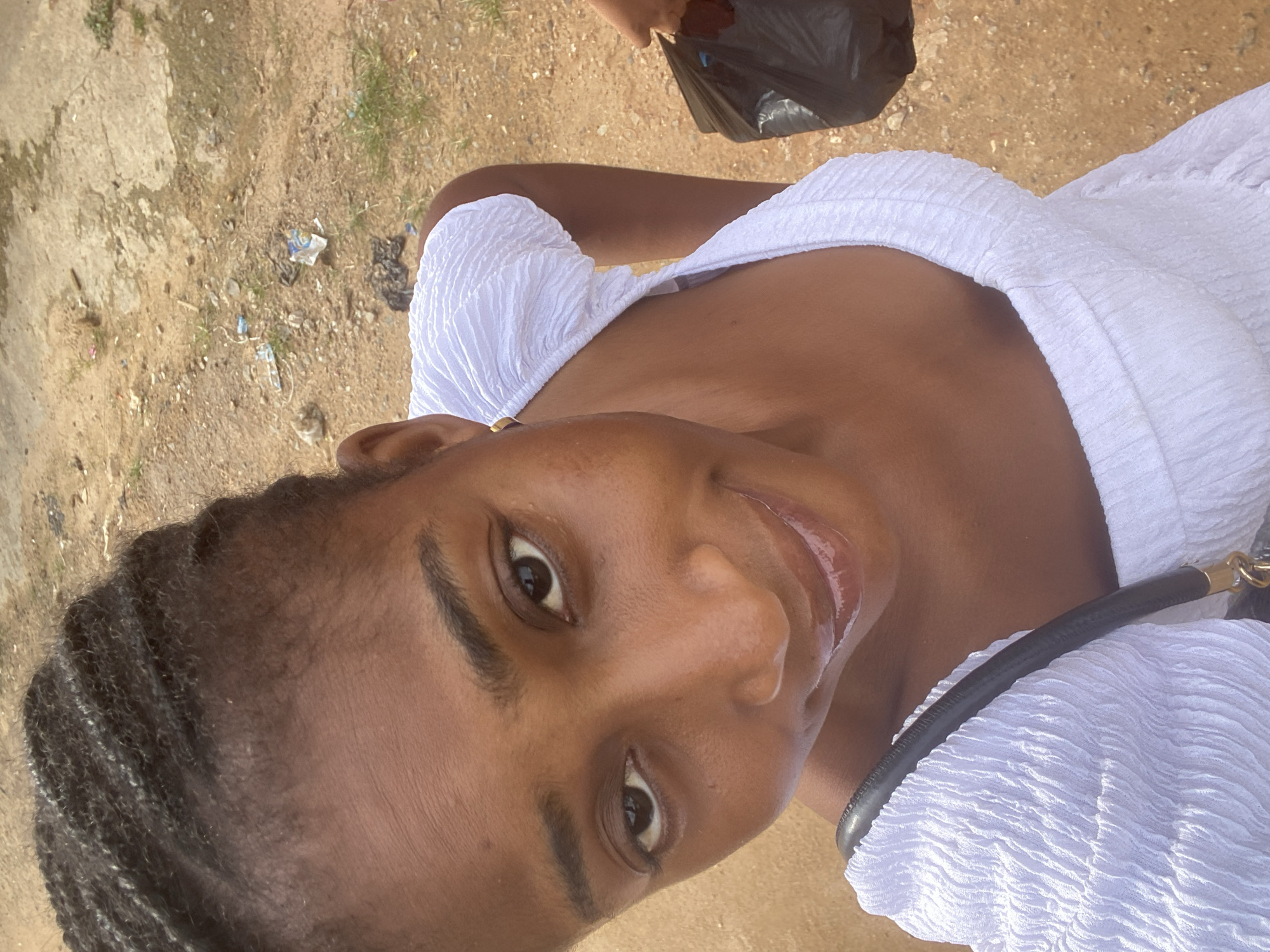 A close-up, high-angle shot captures a young Black woman with braided hair and a white off-the-shoulder top. She is looking up at the camera with a slight smile. Her lips are glossy, and she is wearing subtle makeup. A black purse strap is visible across her chest. In the background, a dirt ground with scattered debris and a person holding a black plastic bag is visible. The scene appears to be outdoors in a possibly urban or semi-urban environment, consistent with a location in Kubwa, Nigeria.