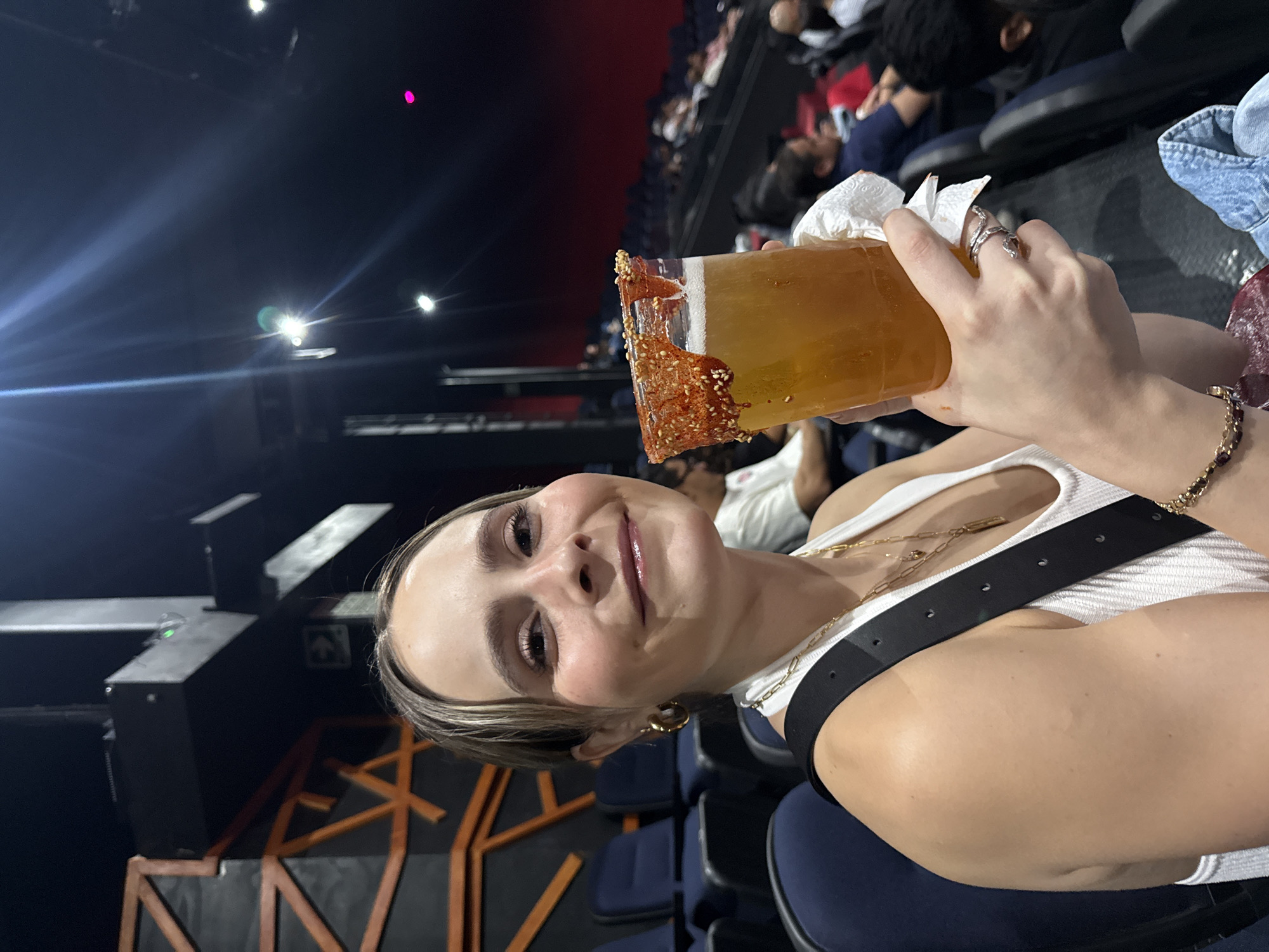A person holds a tall, rimmed glass filled with a yellow beverage, possibly a cocktail, while sitting in a theater-style seating arrangement. The rim of the glass is decorated with a reddish-orange substance and sesame seeds. The person is wearing a white tank top and a black belt with gold accents and has gold hoop earrings and a bracelet. In the background, rows of seats are visible, some occupied by other individuals. The location is Tlalnepantla, Mexico.
