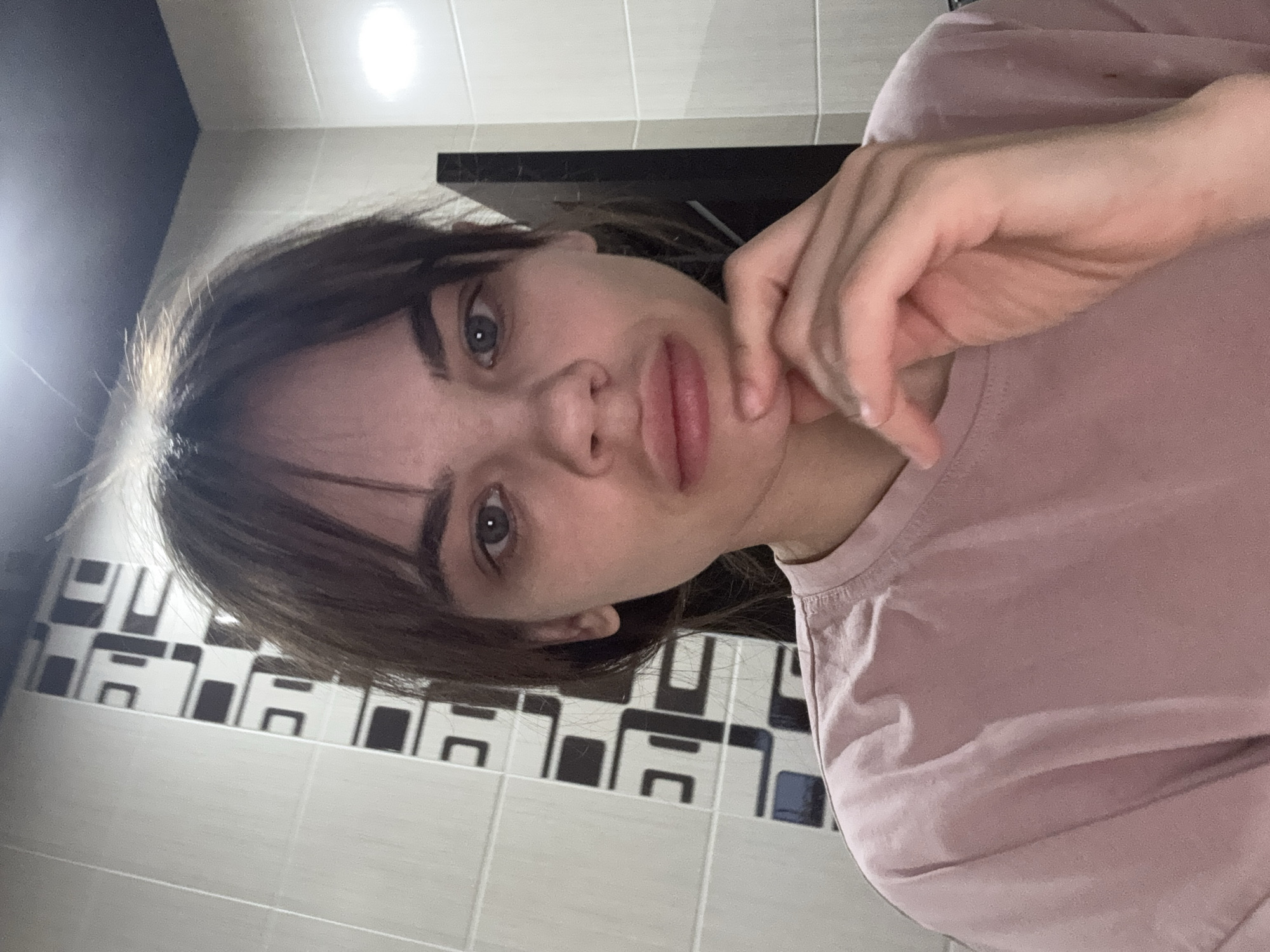 A close-up, rotated selfie shows a young woman, likely in Rostov-on-Don, Russia, looking directly at the camera with a neutral, slightly pensive expression. Her dark hair, styled with bangs, frames her face, highlighting her prominent eyebrows and full lips. She has light-colored eyes, possibly blue or grey. Her right hand is gently resting against her chin or jawline. She is wearing a plain, light pink short-sleeved t-shirt.
The background consists of a wall covered in two sections of tiles: a lower section of light grey or white tiles, and an upper section with a distinct geometric pattern of alternating black and white squares and rectangles. Above the tiled wall, a dark, flat surface, possibly a cabinet or shelf, is visible. The ceiling is light-colored, and a bright, circular artificial light source is visible in the upper part of the frame, illuminating the indoor setting. The scene appears to be an interior space, possibly a kitchen or bathroom, indicated by the tiled walls. There is no visible text.