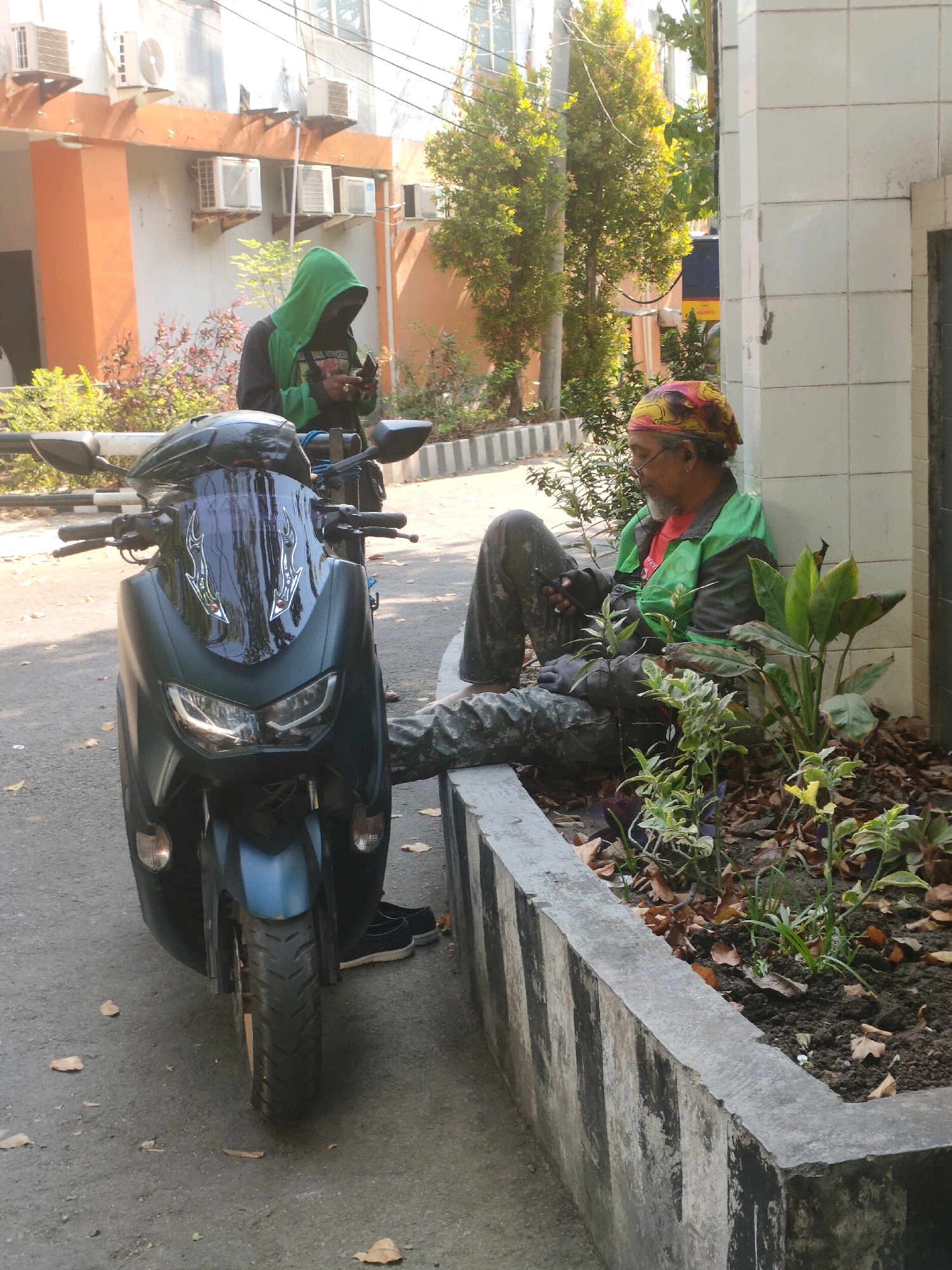 The image captures an outdoor scene near a building in Surabaya, Indonesia, on a sunny day. In the foreground, a dark blue motorcycle is parked on an asphalt road, facing the camera. A person wearing a green hooded jacket and a dark shirt stands behind the motorcycle, looking down at a mobile phone. To the right, leaning against the white tiled wall of the building, a person with a colorful bandana on their head is seated on the ground, also looking at a mobile phone. This individual is wearing a green and red jacket and camouflage pants. In front of the seated person is a planter box filled with soil, fallen leaves, and various plants, bordered by a concrete wall with black and white stripes. The building in the background has orange and white elements, and multiple air conditioning units are visible on its facade. Trees and other greenery are present in the background. The overall atmosphere suggests a casual moment with two individuals engaged with their devices outdoors.