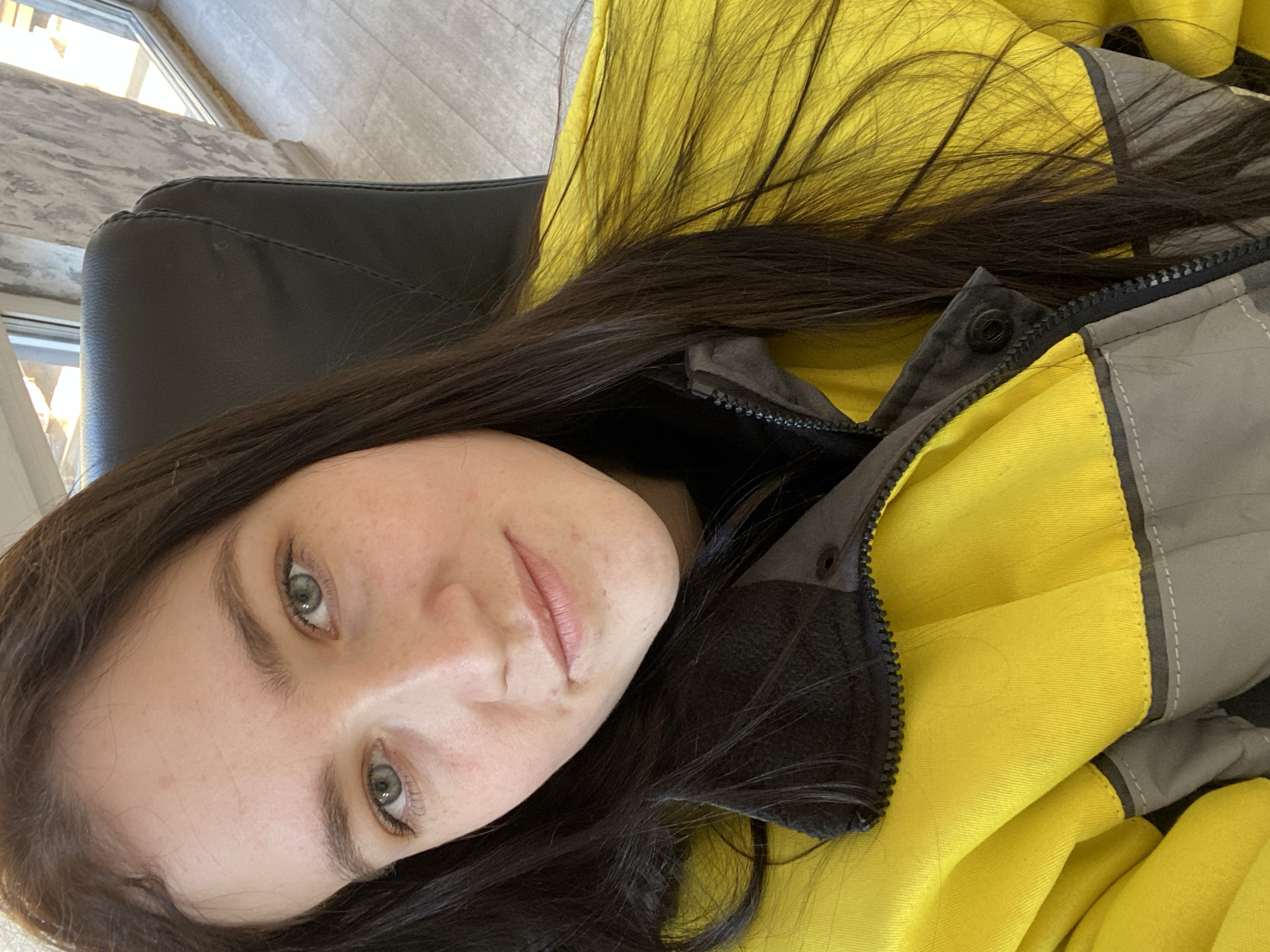 A portrait of a woman with long, dark brown hair and light green eyes is taken indoors. She is wearing a yellow and gray jacket. The woman's face is partially visible, with a slight smile. A black chair is partially visible in the background, and a window lets in natural light. The location of the photo is unknown.
