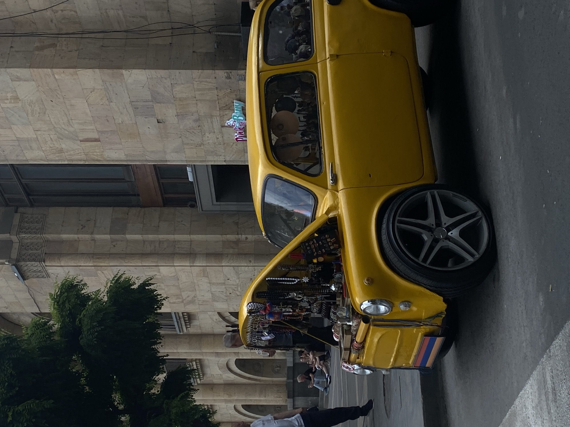 A bright yellow, vintage-style small car, possibly a modified Fiat 600, serves as a mobile vendor stall parked alongside a large stone building in an urban setting. The car's front hood is open, revealing an array of items for sale, including necklaces, bracelets, keychains, and other small souvenirs, meticulously arranged on racks and trays. The car features custom multi-spoke rims on its visible right front wheel and has a stylized license plate on the front bumper displaying the red, blue, and orange colors of the Armenian flag, confirming the location as Yerevan, Armenia.
Several people are present in the scene. One person in a white shirt and dark pants stands near the front of the car, seemingly observing. In the mid-ground, a few individuals are seated on a bench or wall, while others walk or stand casually further back. The overall atmosphere appears relaxed, suggesting a daytime street market or tourist area.
The background features a multi-story building constructed from light-colored stone blocks, exhibiting classical architectural elements such as cornices and recessed windows. Power lines are visible running across the top portion of the image, above the building. A vibrant green tree is partially visible on the left side, adding natural contrast to the urban hardscape. Through the car's side windows, various hats or round objects are discernible inside the vehicle. A light blue and pink sign with partially visible text "Pink B..." is affixed to the building wall in the background. The street is paved with gray asphalt. The time of day appears to be daytime, with clear lighting.