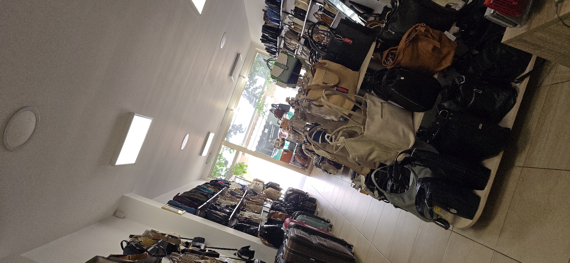 The image depicts the interior of a retail store selling handbags. The store is brightly lit with overhead fluorescent and recessed lighting. Various shelves are packed with handbags of different sizes, colors, and styles. In the foreground, several handbags are displayed on a wooden stand. To the left, luggage and more handbags are visible. The background features a large window and a door, allowing a view of greenery and possibly another building. The floor is tiled. The scene is an indoor commercial space dedicated to the sale of handbags.