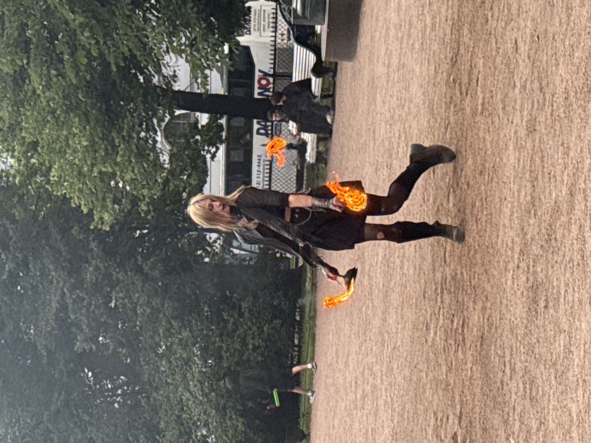This media file shows a street performer in Saint Petersburg, Russia, performing with fire. The subject is a woman with blonde hair, wearing a black outfit and dark boots. She is in motion, with flames emanating from her hands. The background features a tree-lined street with buildings and a fence. Another person, dressed in dark clothing, is visible in the background holding a green object. The ground is a light brown, possibly gravel or dirt. The lighting suggests it is daytime, with an overcast sky. The overall mood appears dynamic and energetic due to the fire performance. Visible text includes a sign with "DA" and numbers that appear to be a phone number, partially obscured.